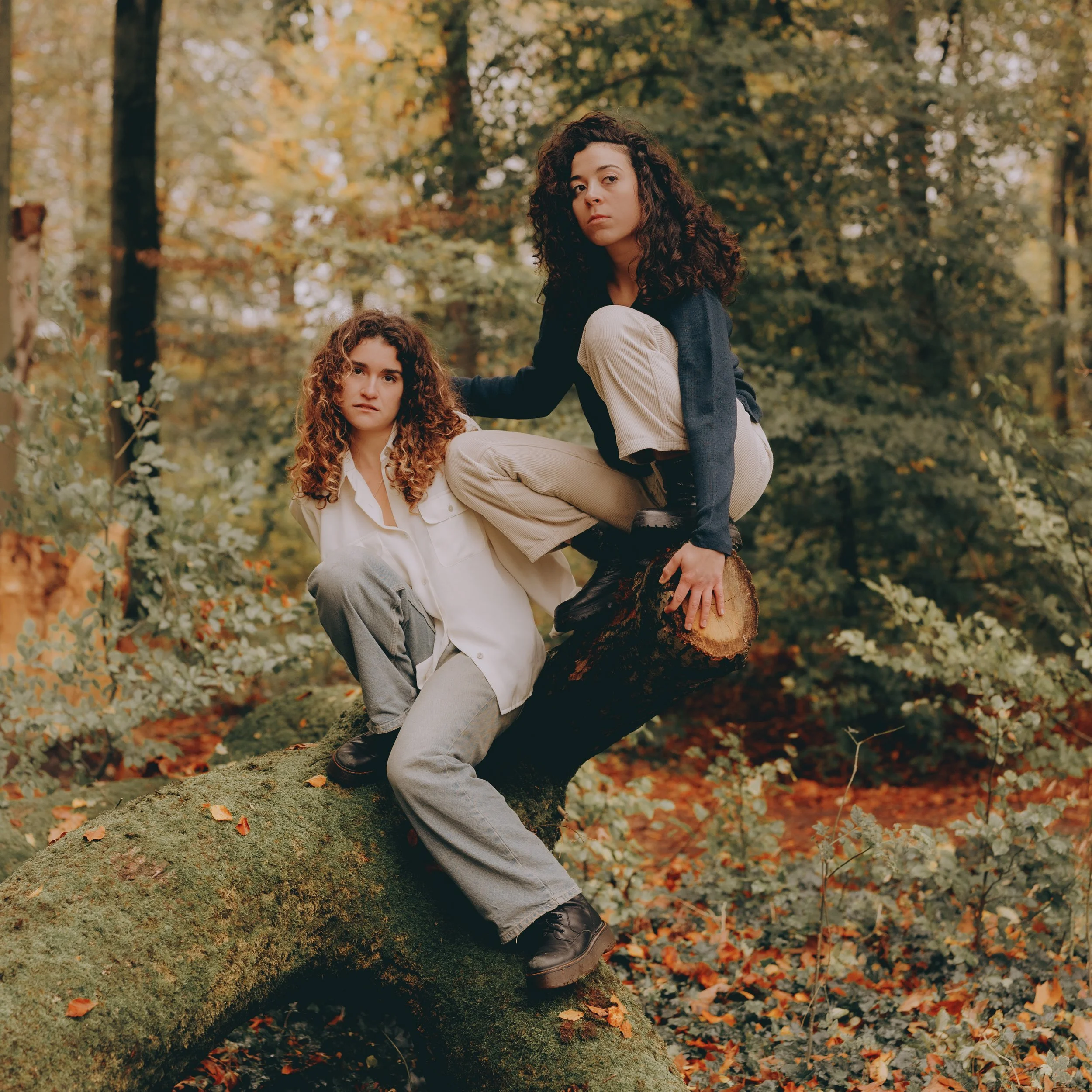 Two women with curly hair climbing and sitting on a fallen tree trunk in a forest with autumn foliage.
