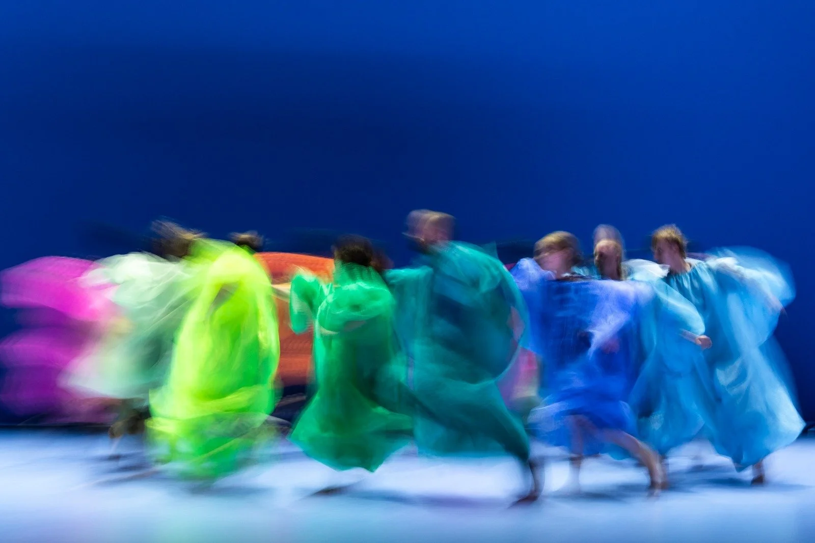 Blurred image of dancers in colorful costumes performing on stage with a blue background.