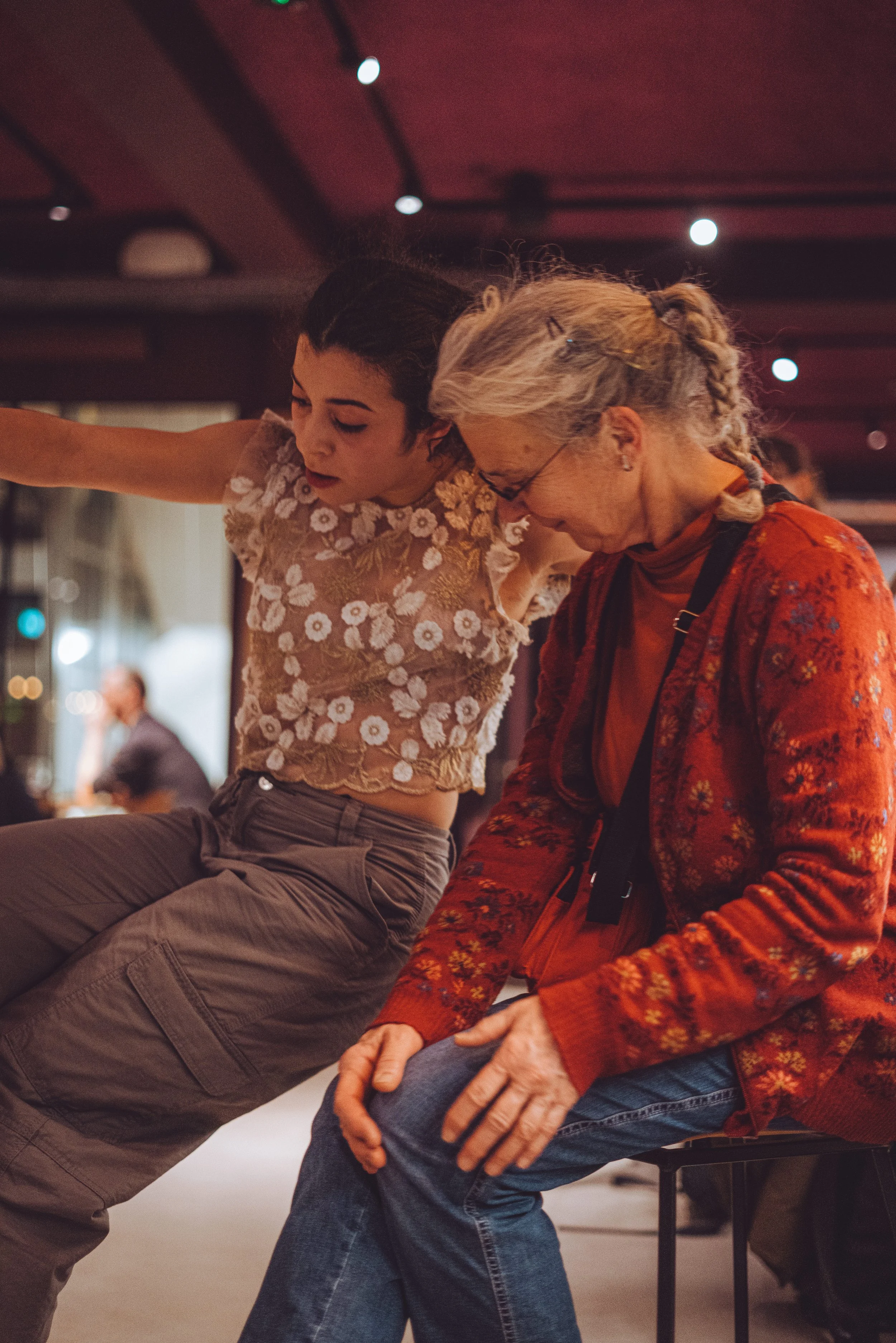 A young woman with dark hair and a floral top leaning towards an elderly woman with gray hair and glasses sitting on a bench, in an indoor setting with warm lighting.