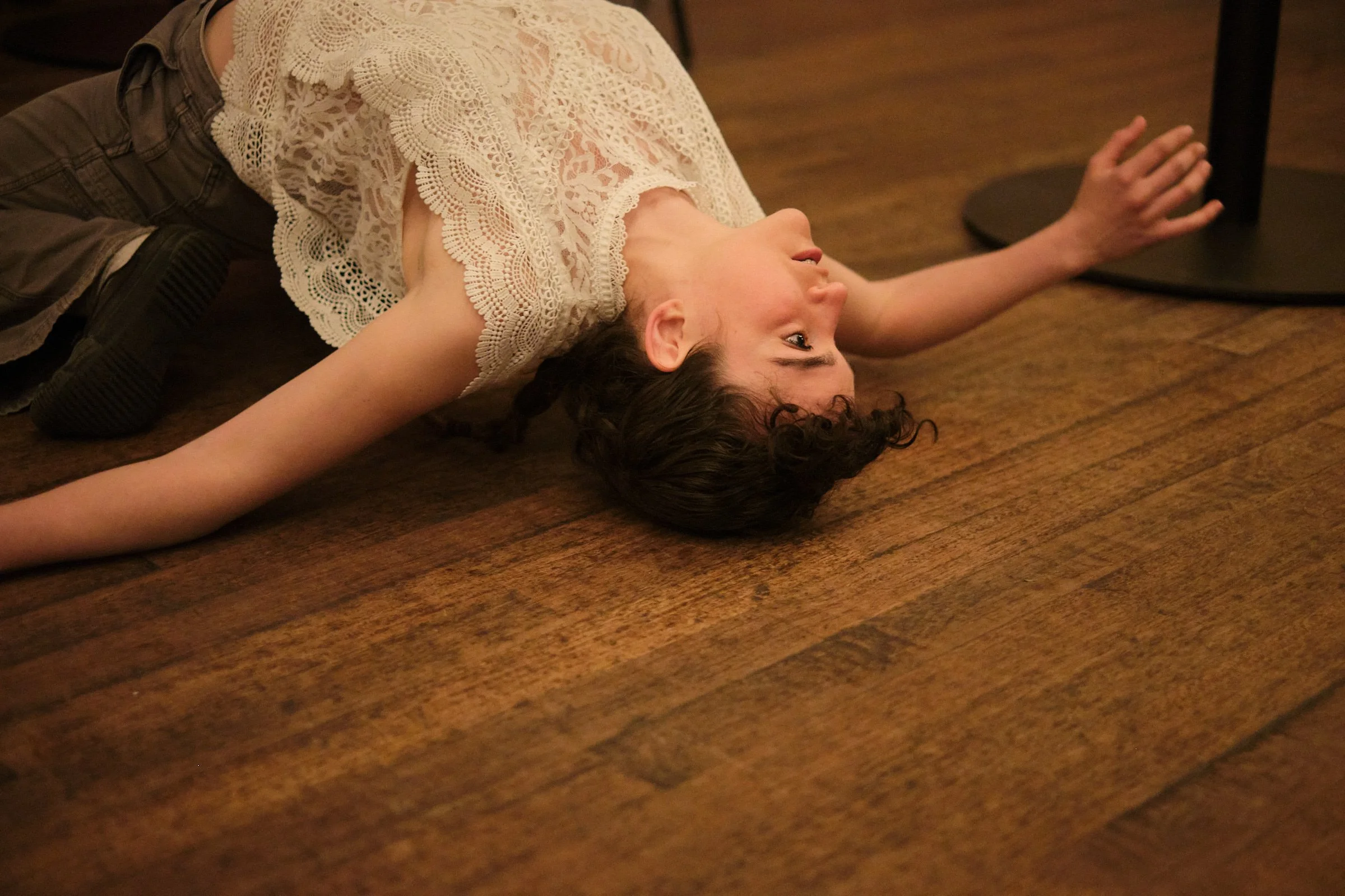 A person lying on their back on a wooden floor with an arm extended and face turned to the side, wearing a lacy white top and brown pants.