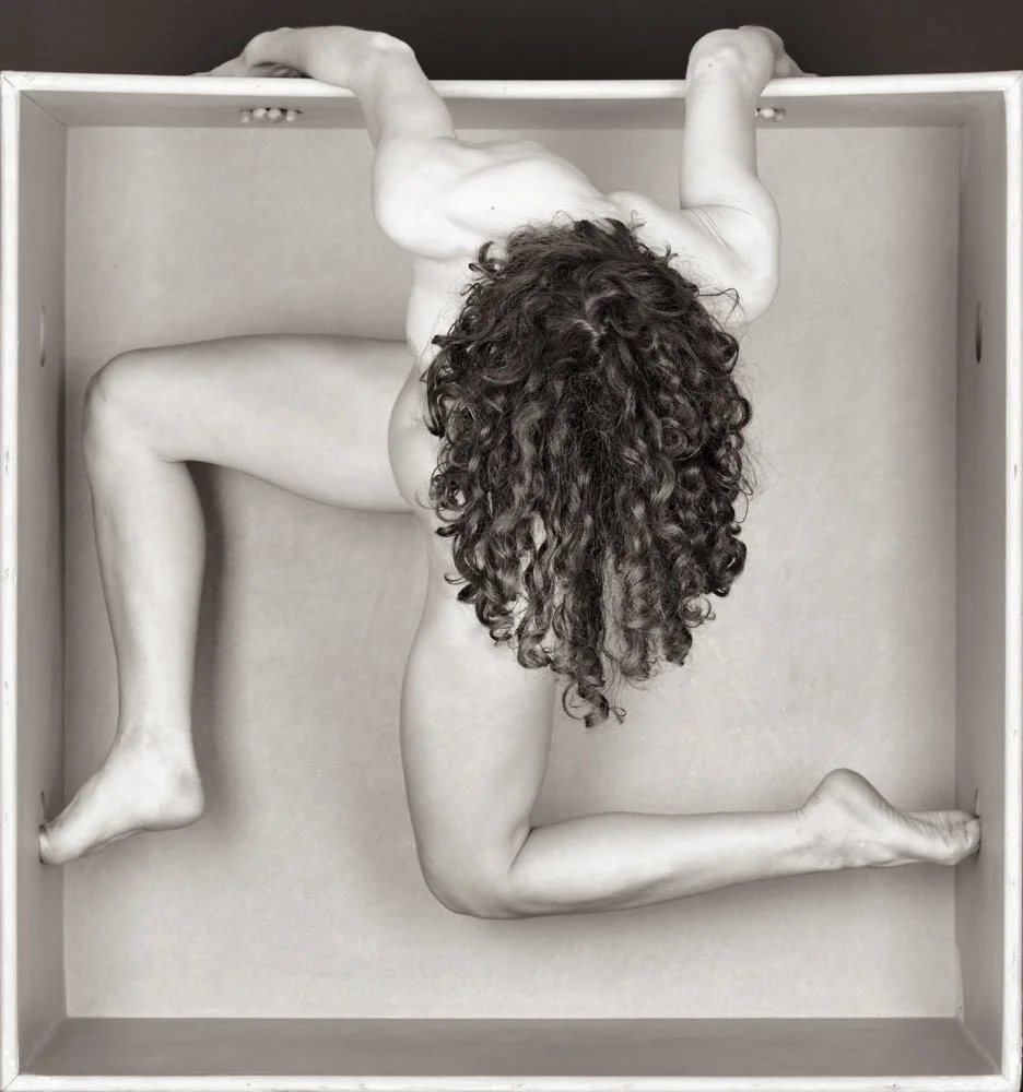 A black and white photo of a woman with curly hair in a box, positioned in a sculptural pose with her head hanging downward, one arm gripping the side of the box and the other bent behind her. The image emphasizes her body and hair in an artistic com