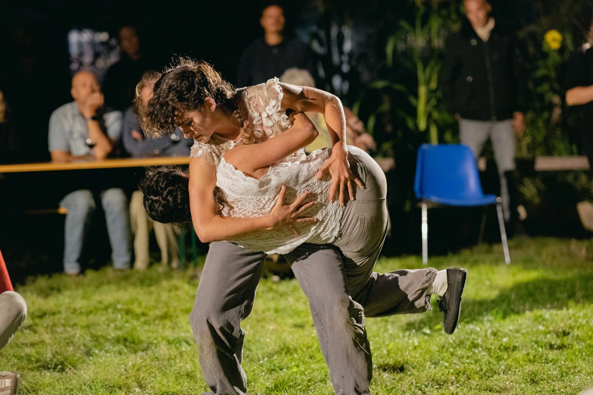 Two people dancing closely at an outdoor event at night, with onlookers seated in the background.