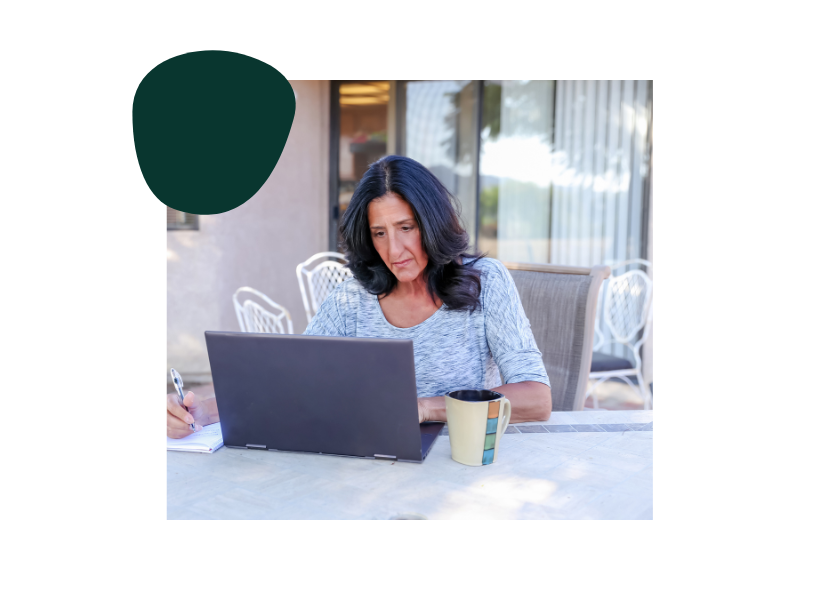 A woman working on a laptop at an outdoor table, with a notebook and pen in hand, and a coffee mug nearby.