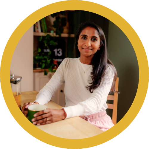 A woman sitting at a kitchen table holding a mug, smiling at the camera.
