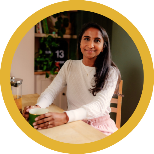 A woman sitting at a kitchen table holding a mug, smiling at the camera, with kitchen shelves and plants in the background.