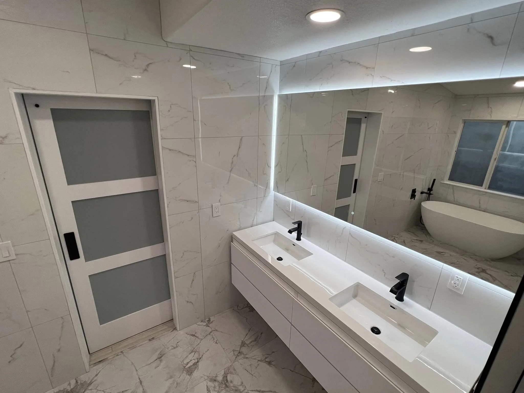 Modern bathroom with white marble walls and floor, double vanity with two rectangular sinks, black faucets, large mirror, and white cabinets underneath. There are LED lights around the mirror and a window with frosted glass.