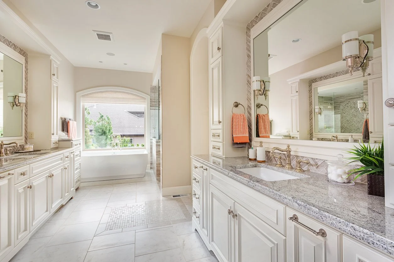 Bright bathroom with white cabinets, granite countertops, large mirror, and a bathtub near a window with outdoor view.