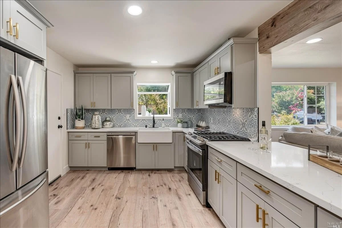 Modern kitchen with light gray cabinets, stainless steel appliances, patterned tile backsplash, white farmhouse sink, and a window overlooking a backyard. The floor is light wood, and there are decorative items on the counter.