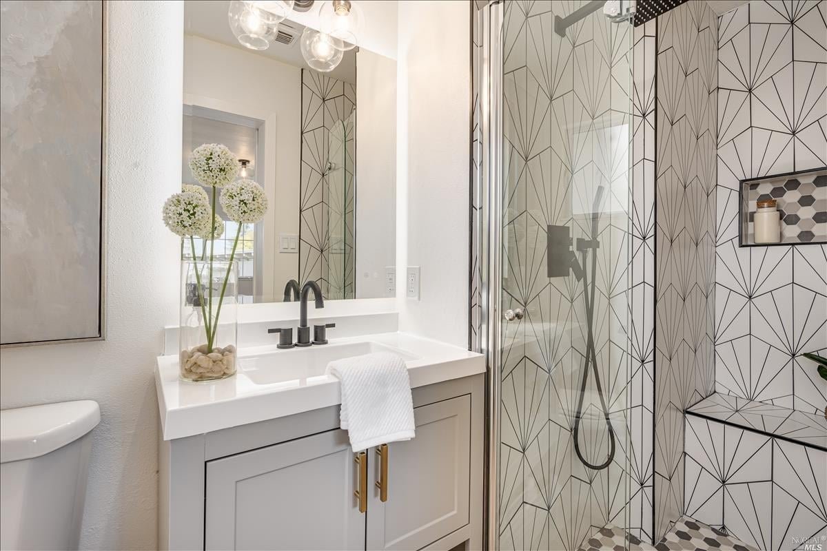Modern bathroom with geometric patterned tiled shower stall, white sink vanity with black fixtures, a large mirror, a vase with white flowers on the vanity, and a toilet visible on the left side.