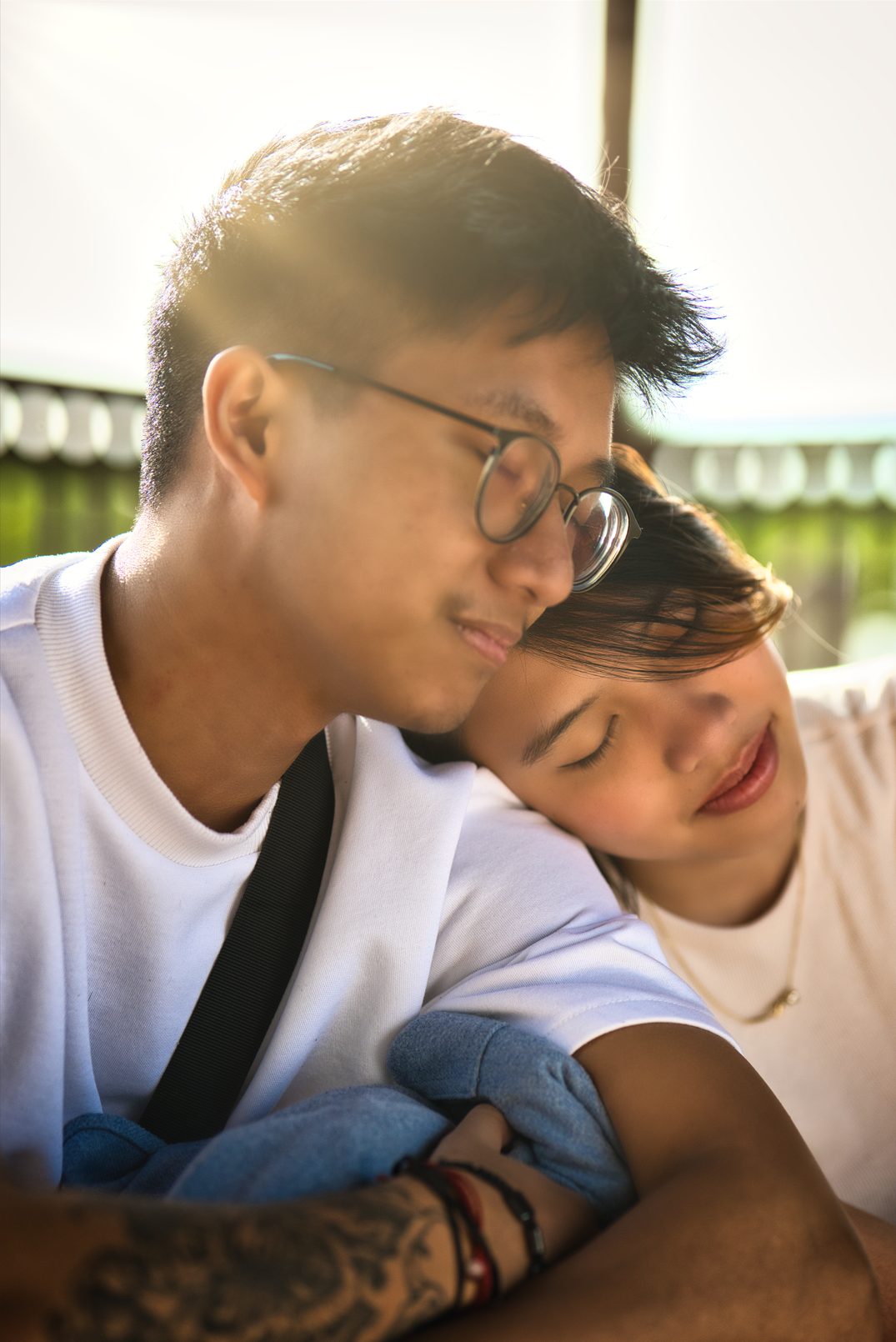 A young man with glasses and short dark hair resting his head on a young woman's shoulder, with her eyes closed, outdoors with sunlight filtering through trees.