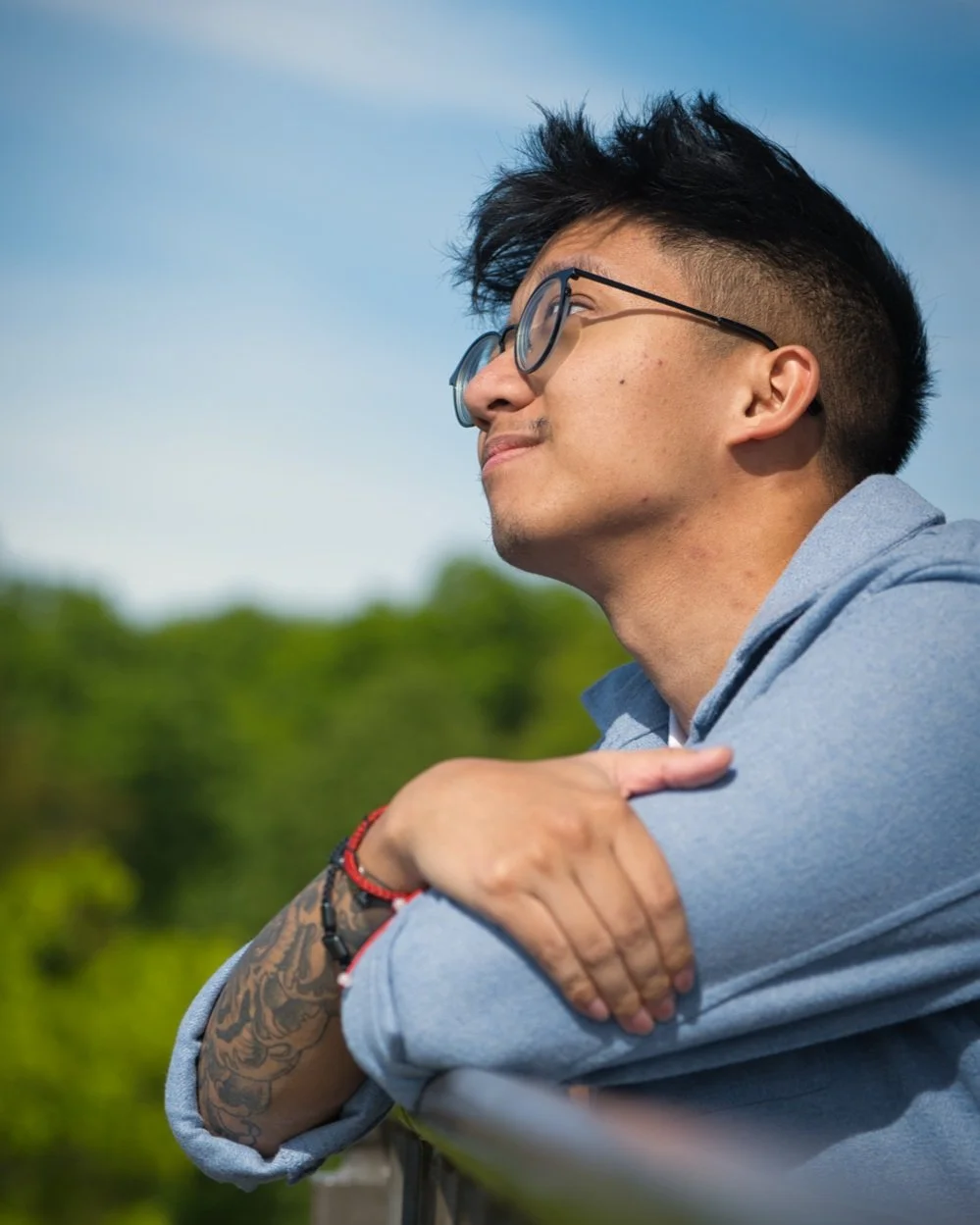 Young man with glasses and tattoos on his arm, smiling and looking into the distance outdoors on a sunny day.