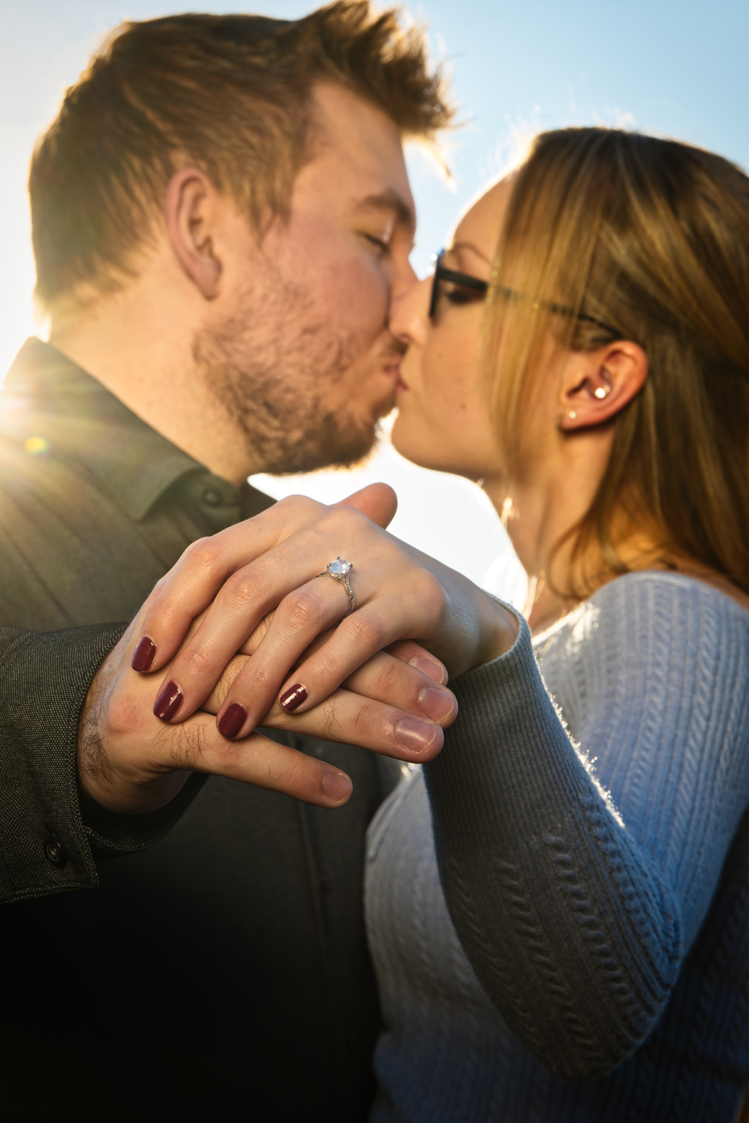 A man and woman kissing outdoors with sunlight in the background. The woman is wearing glasses and has an engagement ring on her finger, and they are holding hands.