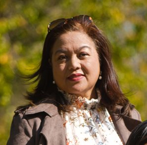 A woman with shoulder-length dark hair, wearing a brown jacket over a floral blouse, outdoors with green foliage in the background.