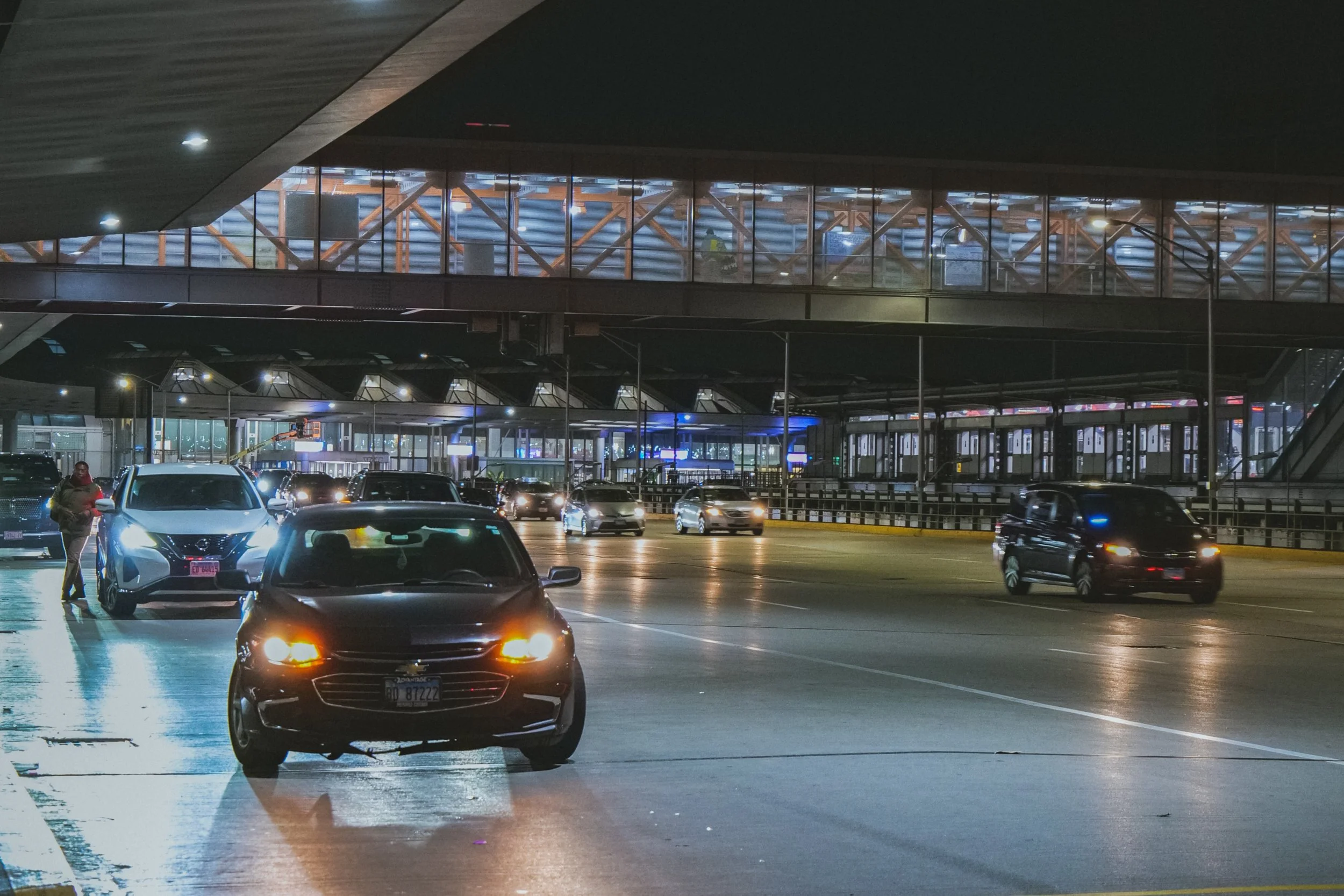 Nighttime scene at an airport terminal with cars parked and driving, an individual standing near a white car, illuminated building structures, and a skybridge connecting to the terminal.