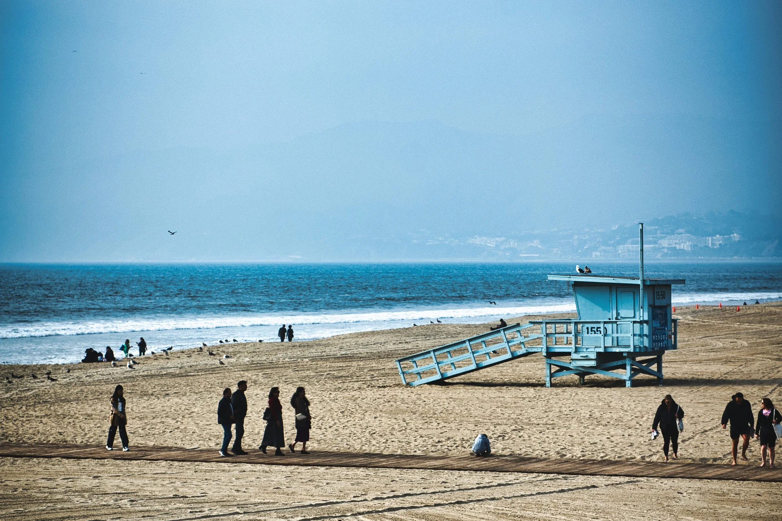 People walking along the sandy beach near a blue lifeguard tower with a ramp, with the ocean and distant shore in the background.