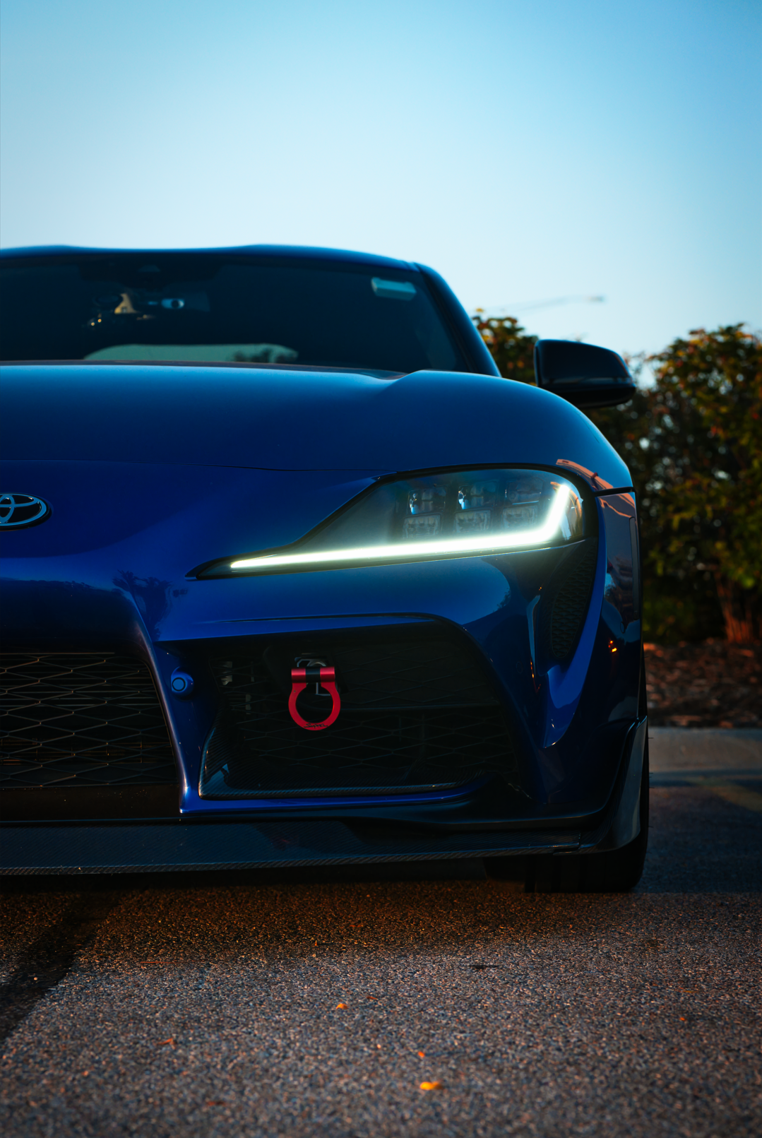 Close-up of the front of a dark blue Toyota sports car with a red tow hook, daytime with a clear blue sky and trees in the background.