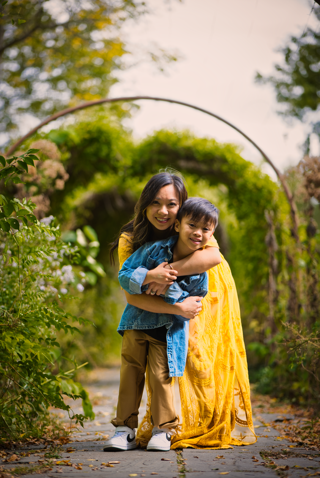 A woman in a yellow dress and a young boy in casual clothing hugging on a tree-lined pathway.