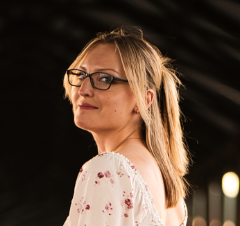 A woman with glasses and blonde hair smiling in a dimly lit indoor setting.