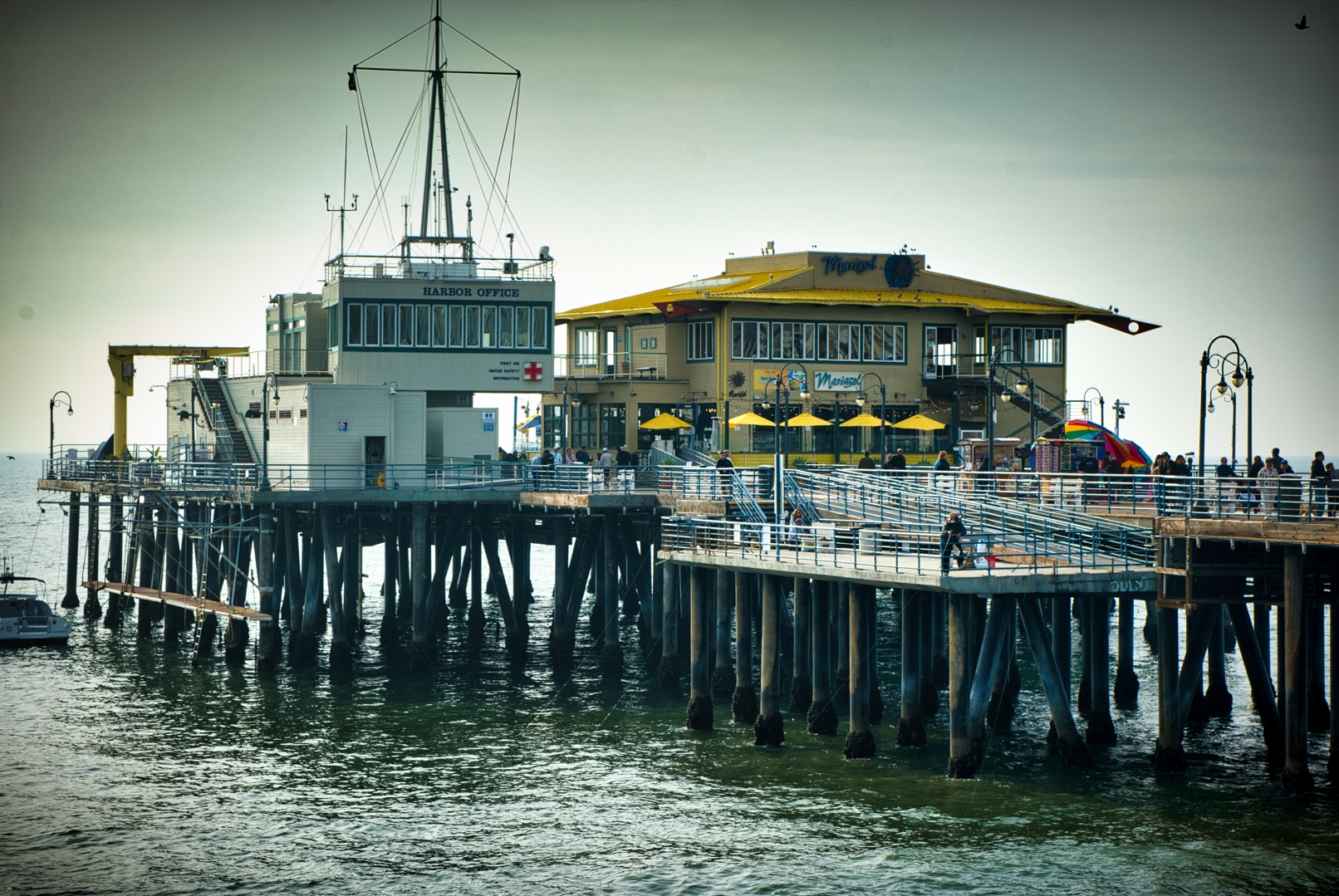 A seaside pier with buildings on stilts over the water, featuring a harbor office, a yellow building with yellow umbrellas, and people walking along the pier.