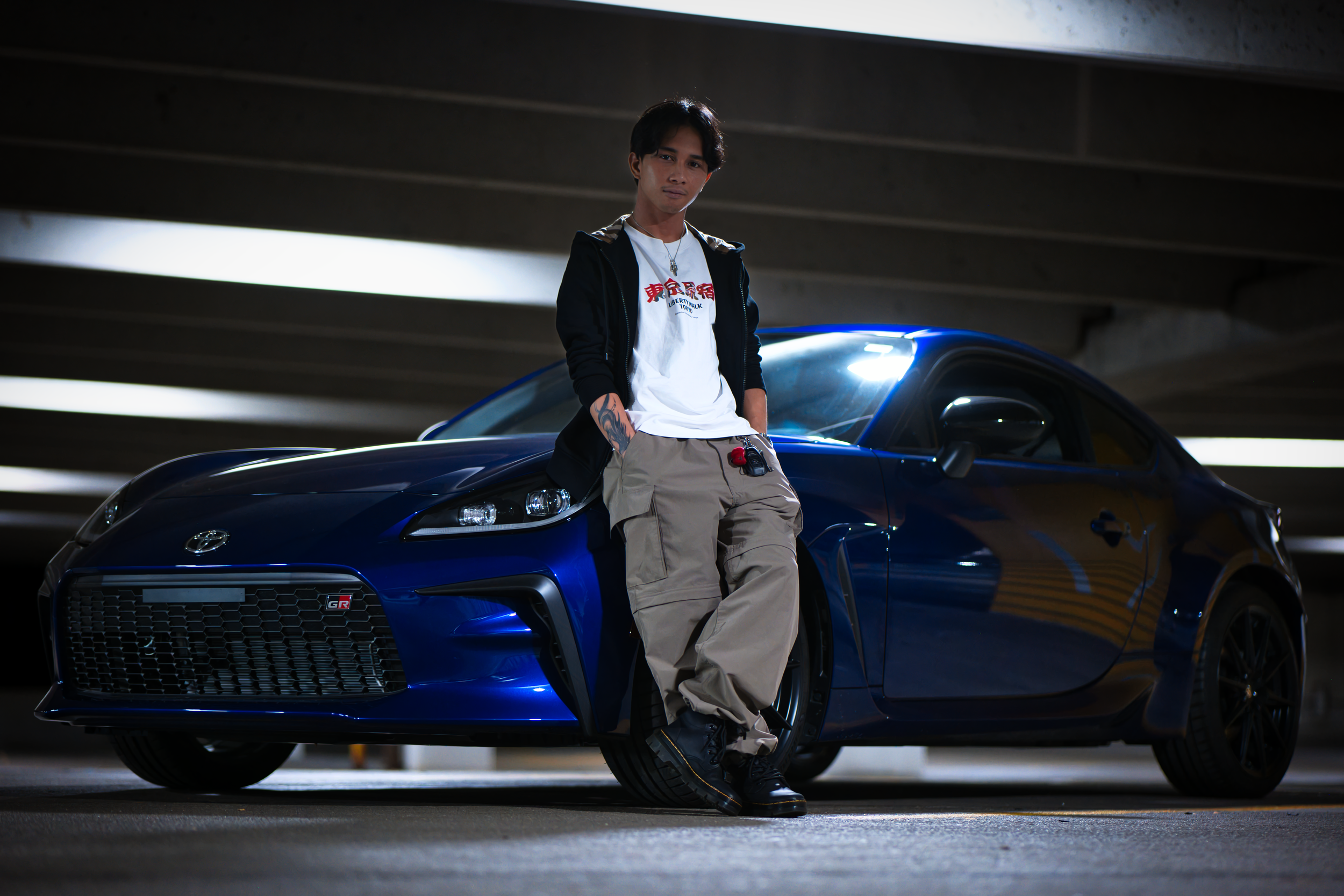 A young man leaning against a blue sports car in an underground parking garage.