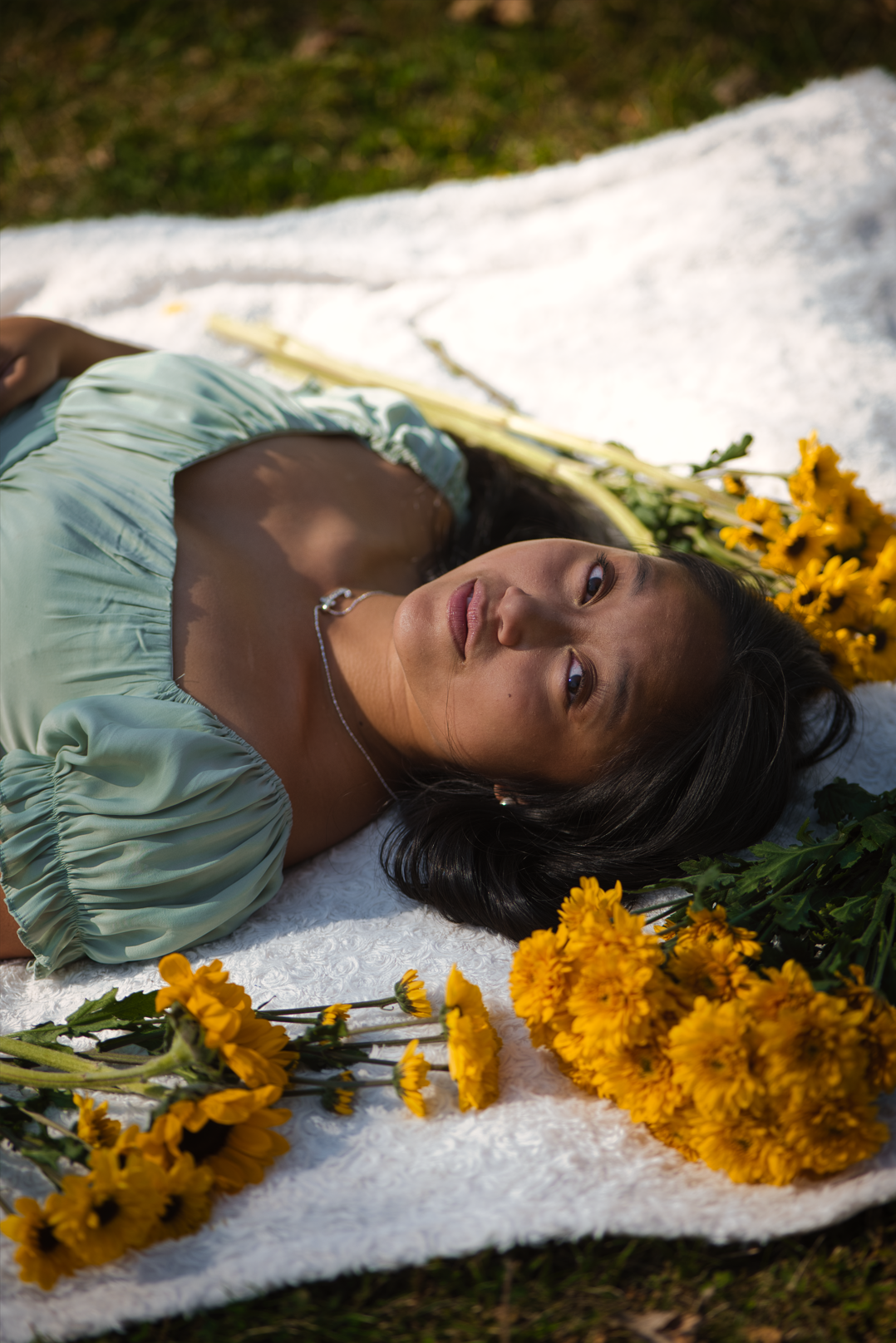A young woman with long black hair lies on a white blanket outdoors surrounded by yellow flowers, looking up with a calm expression.