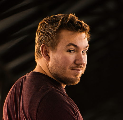 A young man with light skin, short light brown hair, and a beard, looking over his shoulder with a slight smile, wearing a dark maroon shirt.