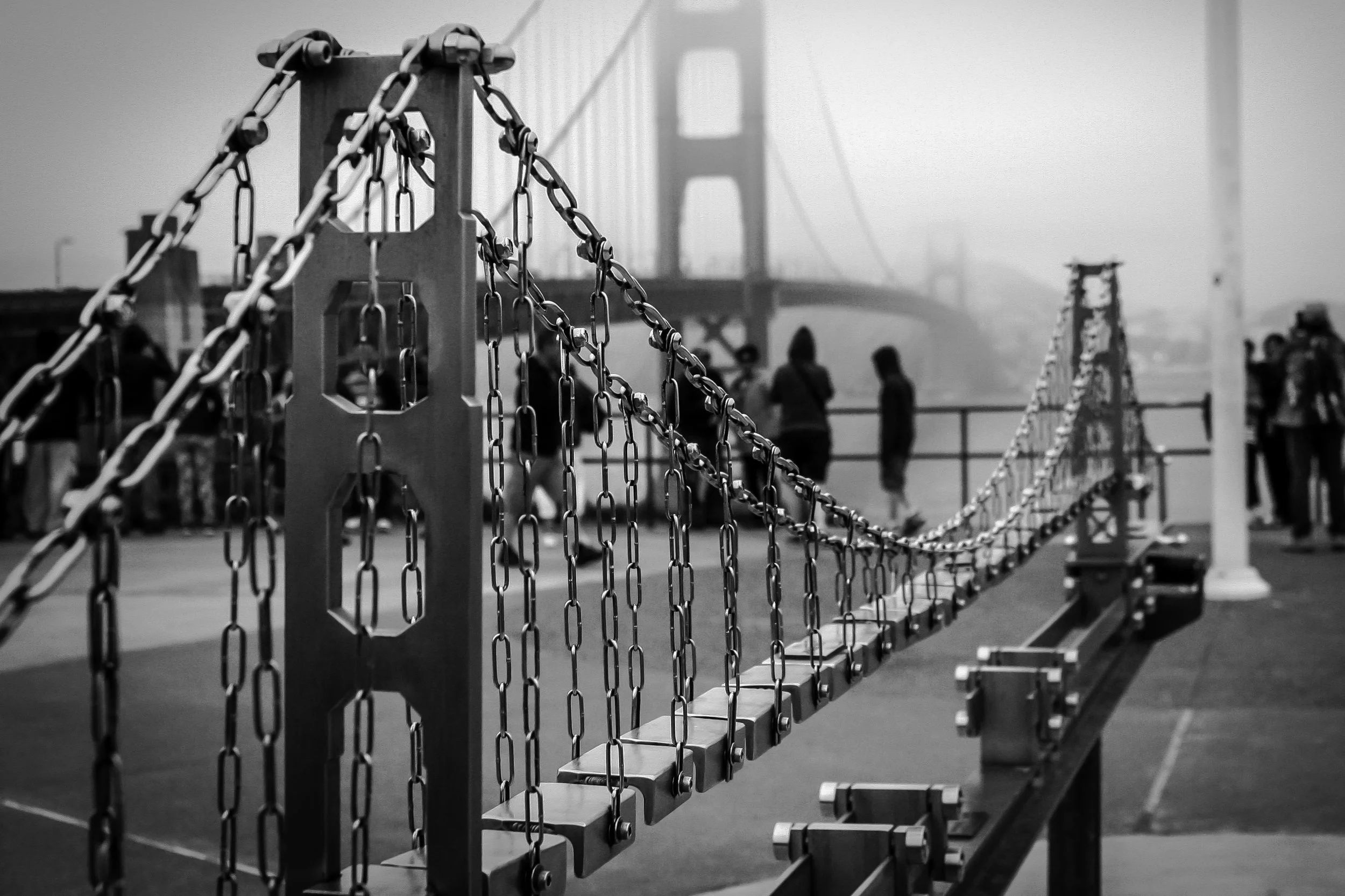 Black-and-white photo of a chain-link fence with a bridge in the background and people walking along a pier or promenade.