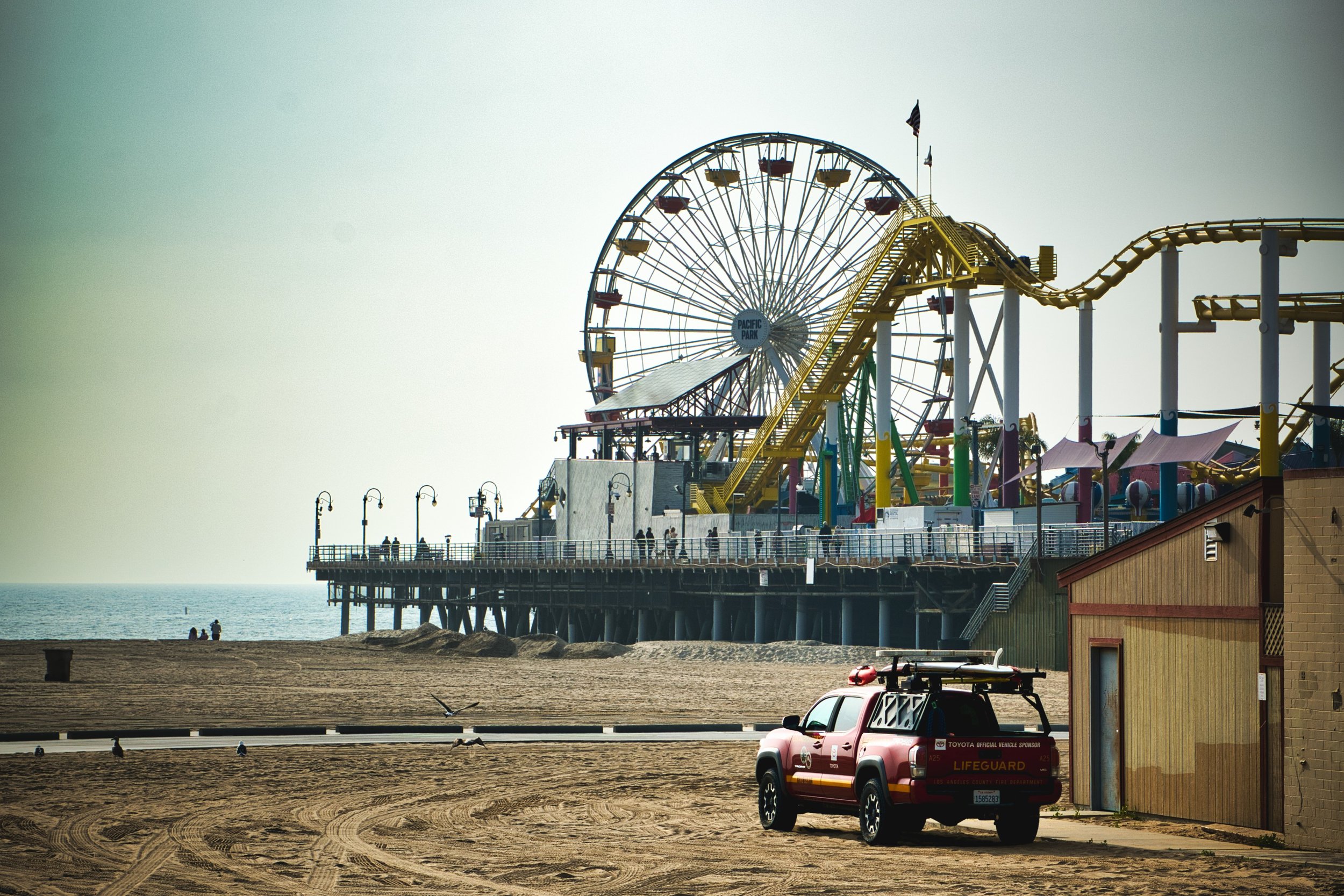 A red lifeguard vehicle parked on sand near a wooden building on a pier with an amusement park, including a large Ferris wheel and roller coaster, in the background at the beach.