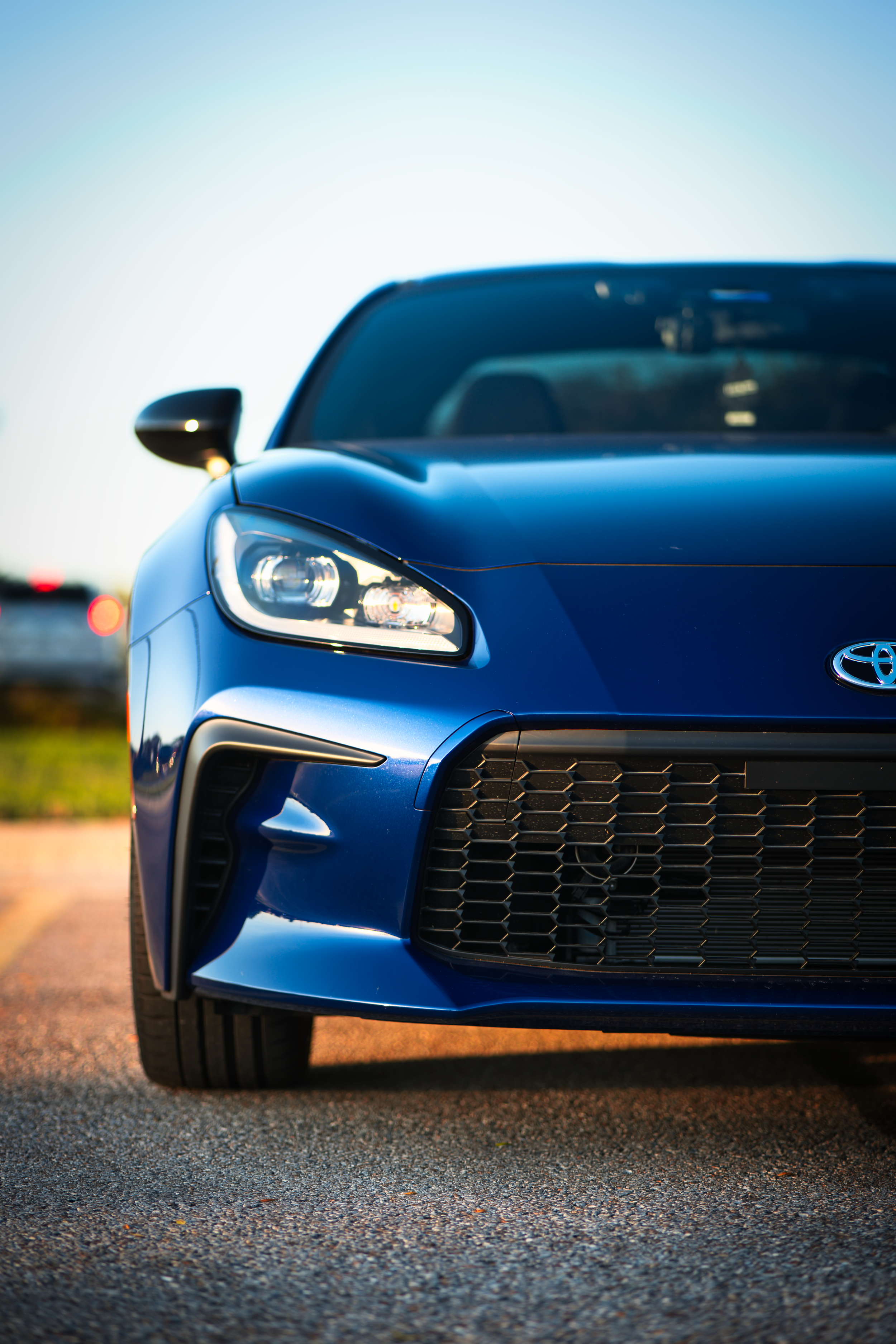 Close-up of the front of a blue Toyota sports car parked on a road with a blurred background of trees and other vehicles.