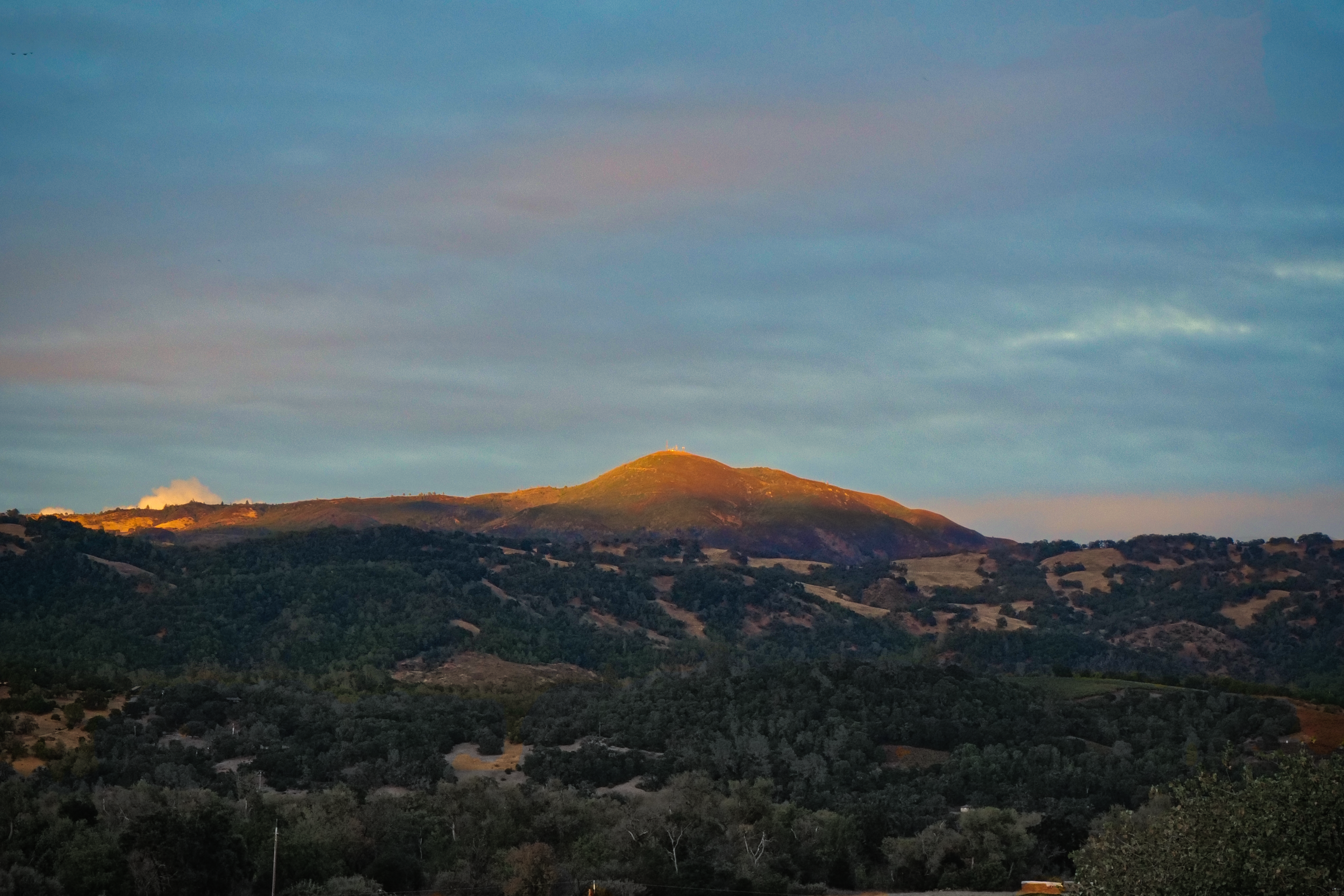 Sunlit hilltop with a small structure or antenna on top, surrounded by rolling hills and trees under a cloudy sky at sunset.