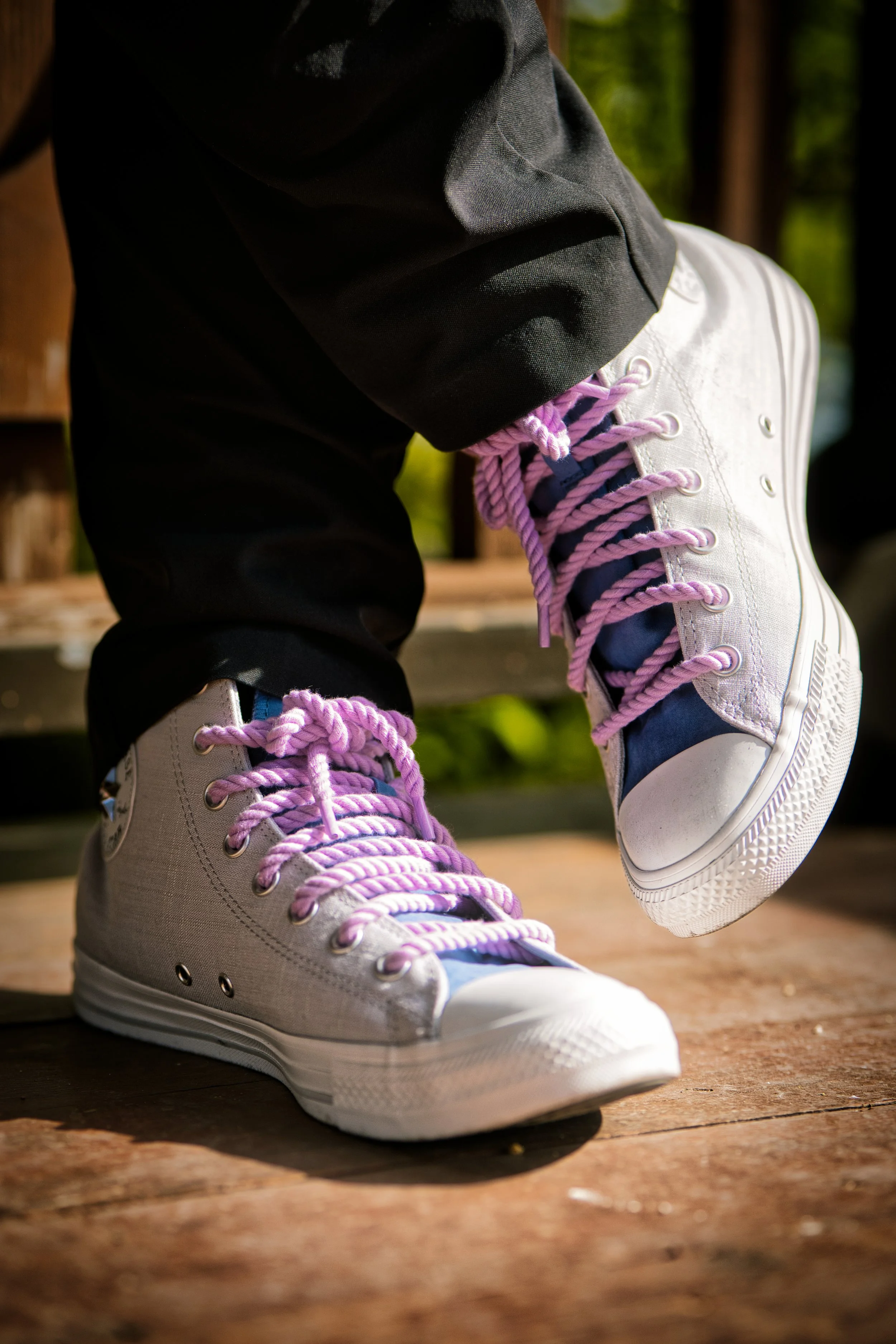Close-up of a person wearing white high-top sneakers with pink laces, black pants, and standing on a wooden surface outdoors.