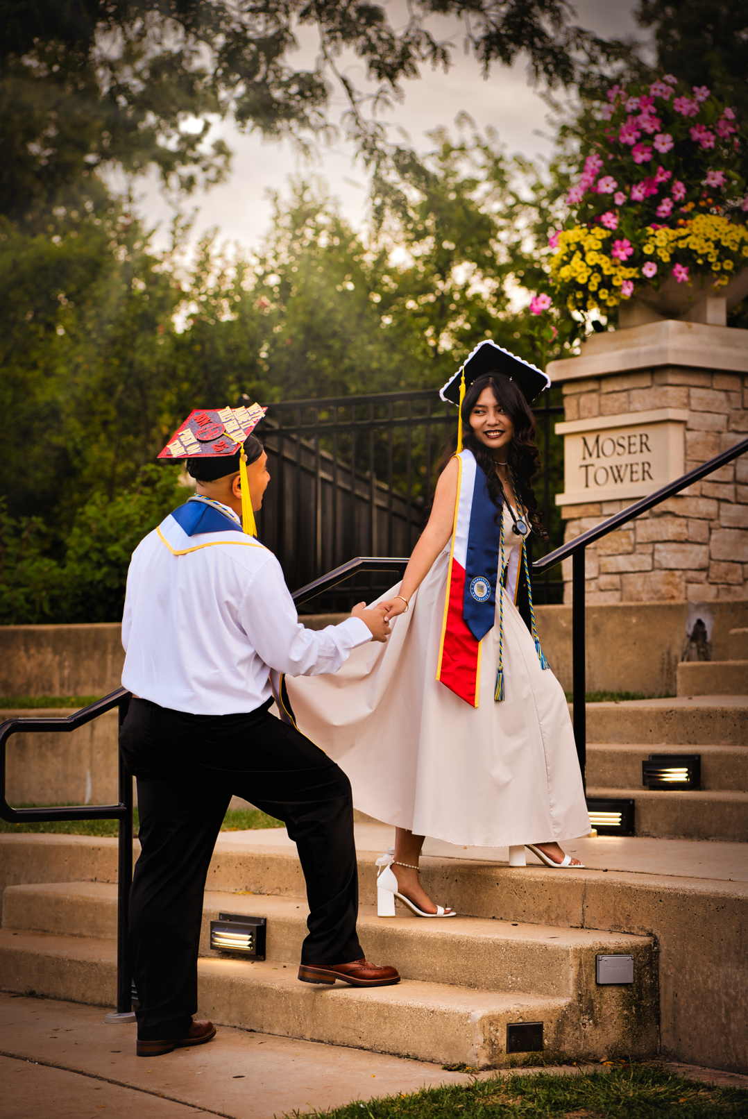 A woman in a graduation gown and cap climbing stairs, holding hands with a man in a white shirt and paper graduation cap, during sunset outside Moser Tower surrounded by greenery and flowers.