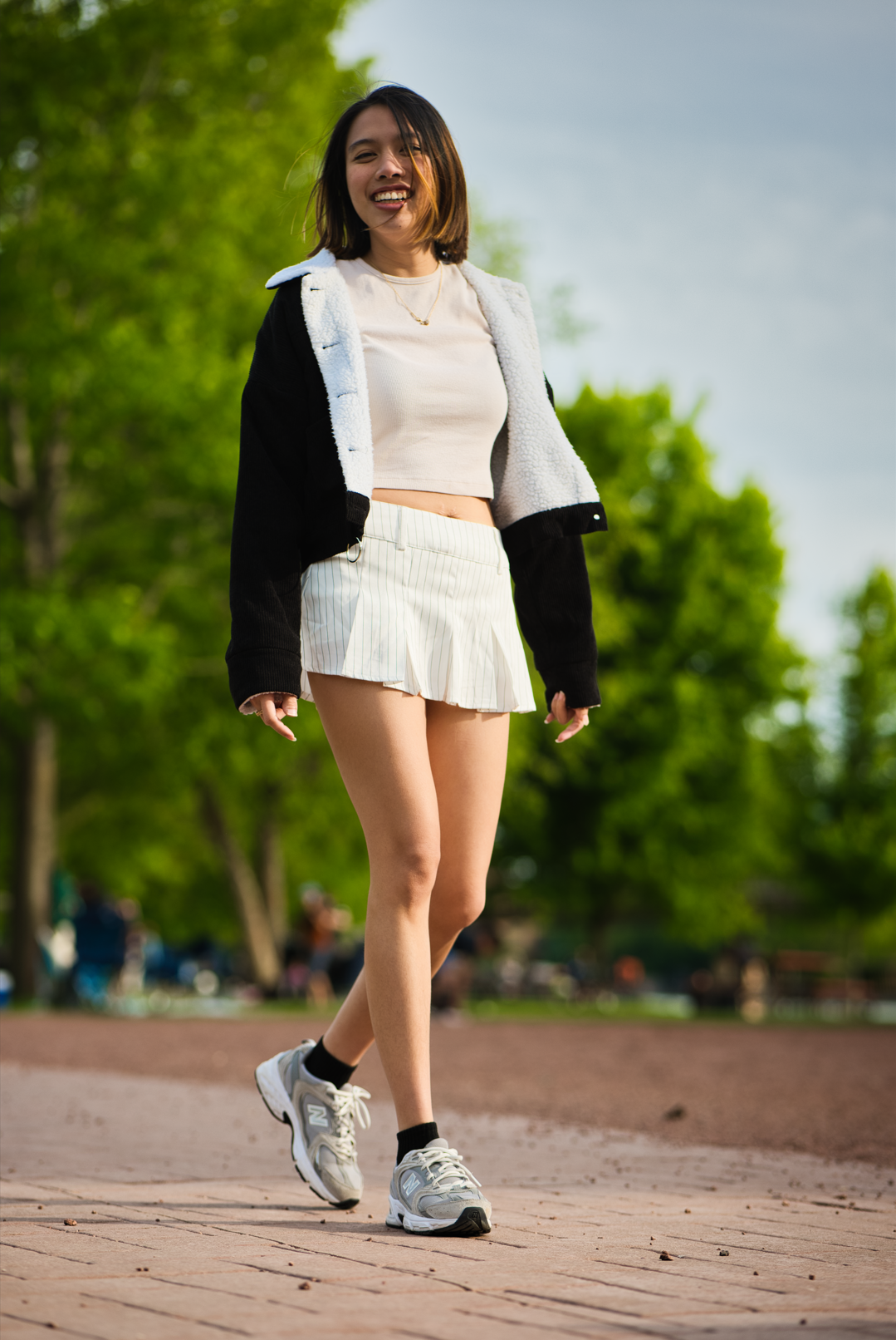 Young woman smiling while walking outdoors in a park, wearing a black and white jacket, white crop top, white pleated skirt, black socks, and gray athletic shoes.
