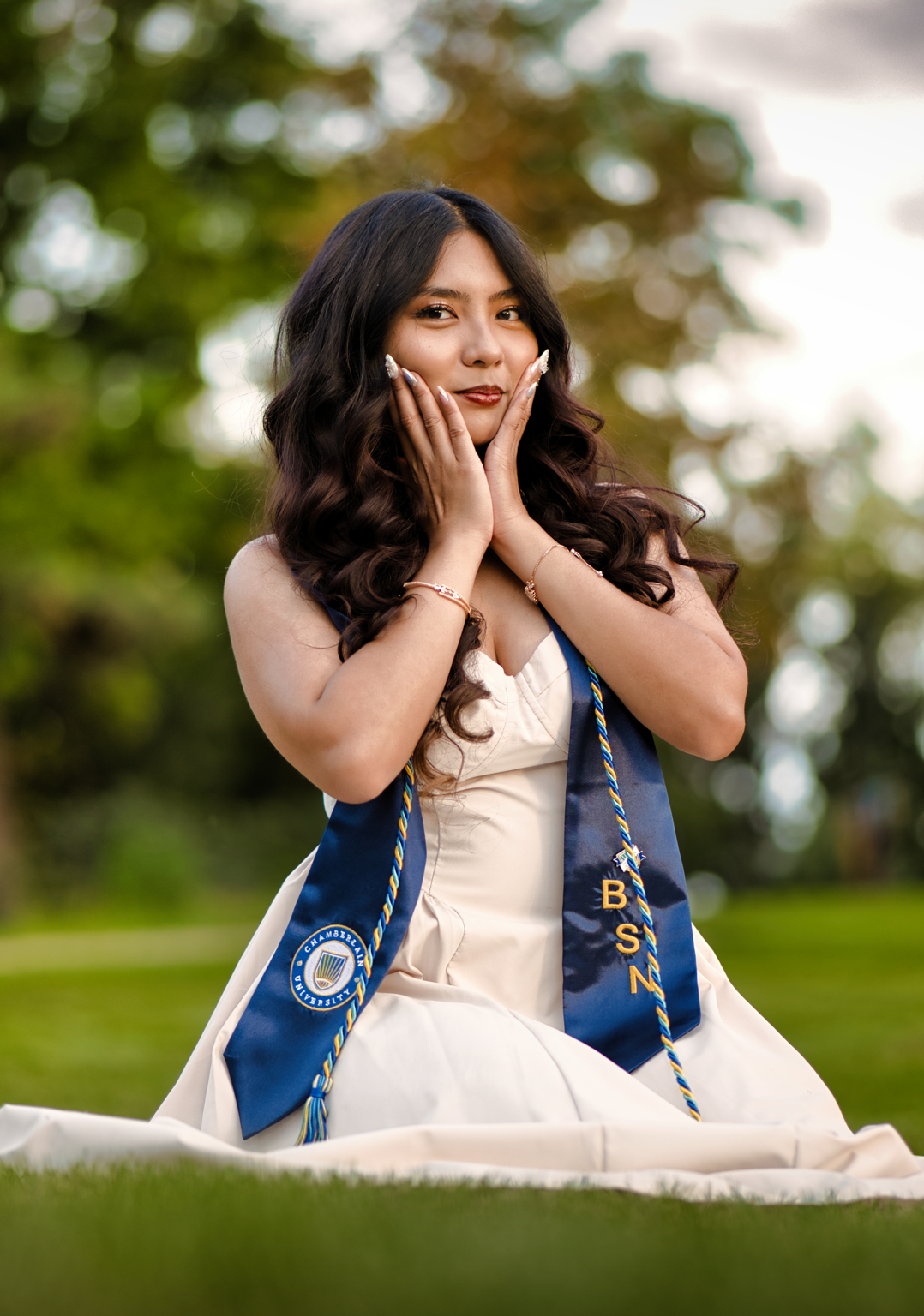 Graduate woman with a diploma and university sash kneeling on grass outdoors, smiling with hands on her cheeks.