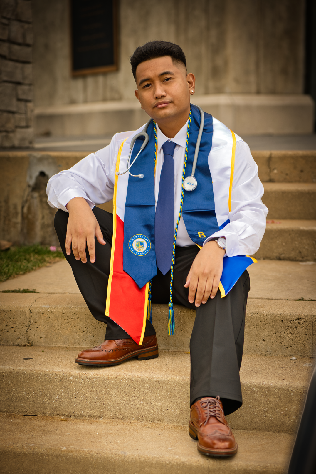 A young man dressed as a doctor, wearing a white coat, blue tie, and a blue and red scarf with the university crest, seated on outdoor stairs. He has a stethoscope around his neck and is looking confidently at the camera.