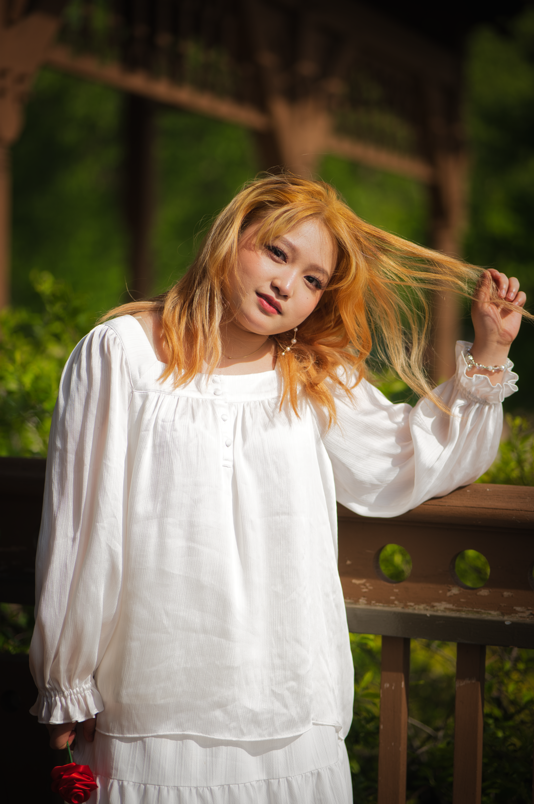 A woman with strawberry blonde hair wearing a white blouse standing outdoors on a wooden bridge, holding a red rose.