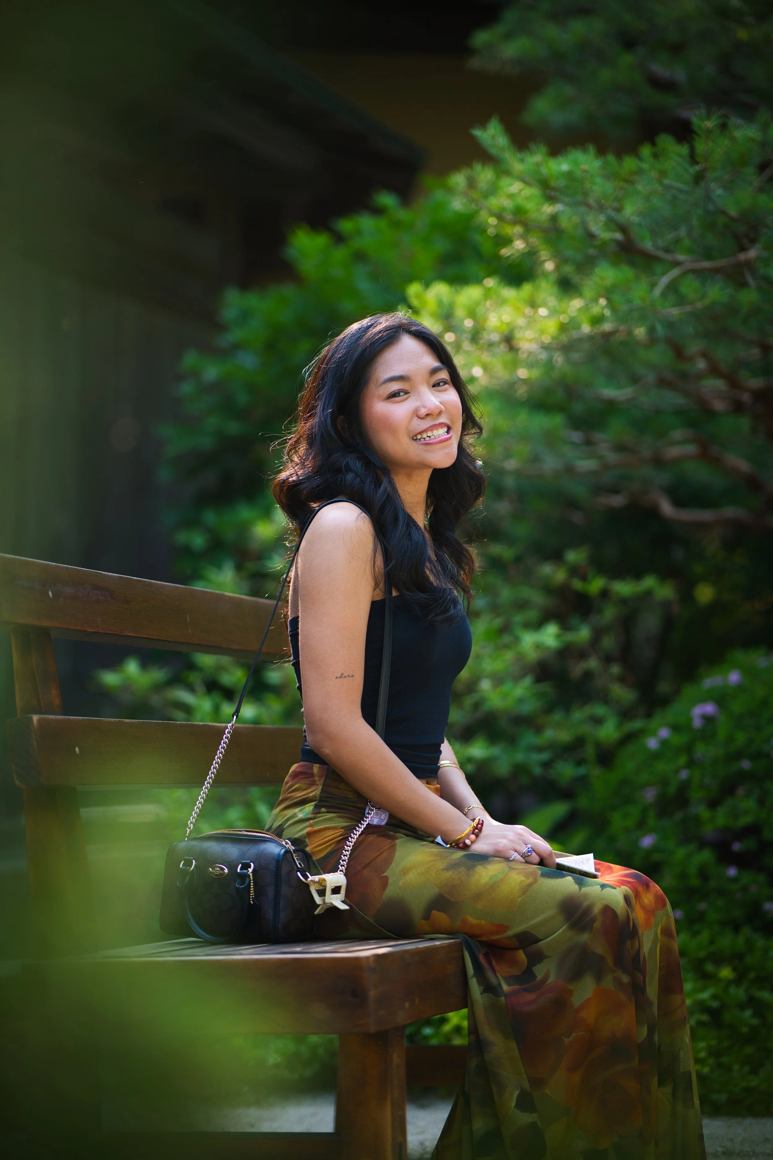 A woman with long black hair sitting on a wooden bench outdoors surrounded by greenery. She is smiling and wearing a black sleeveless top and colorful, patterned skirt, with a small black handbag beside her.