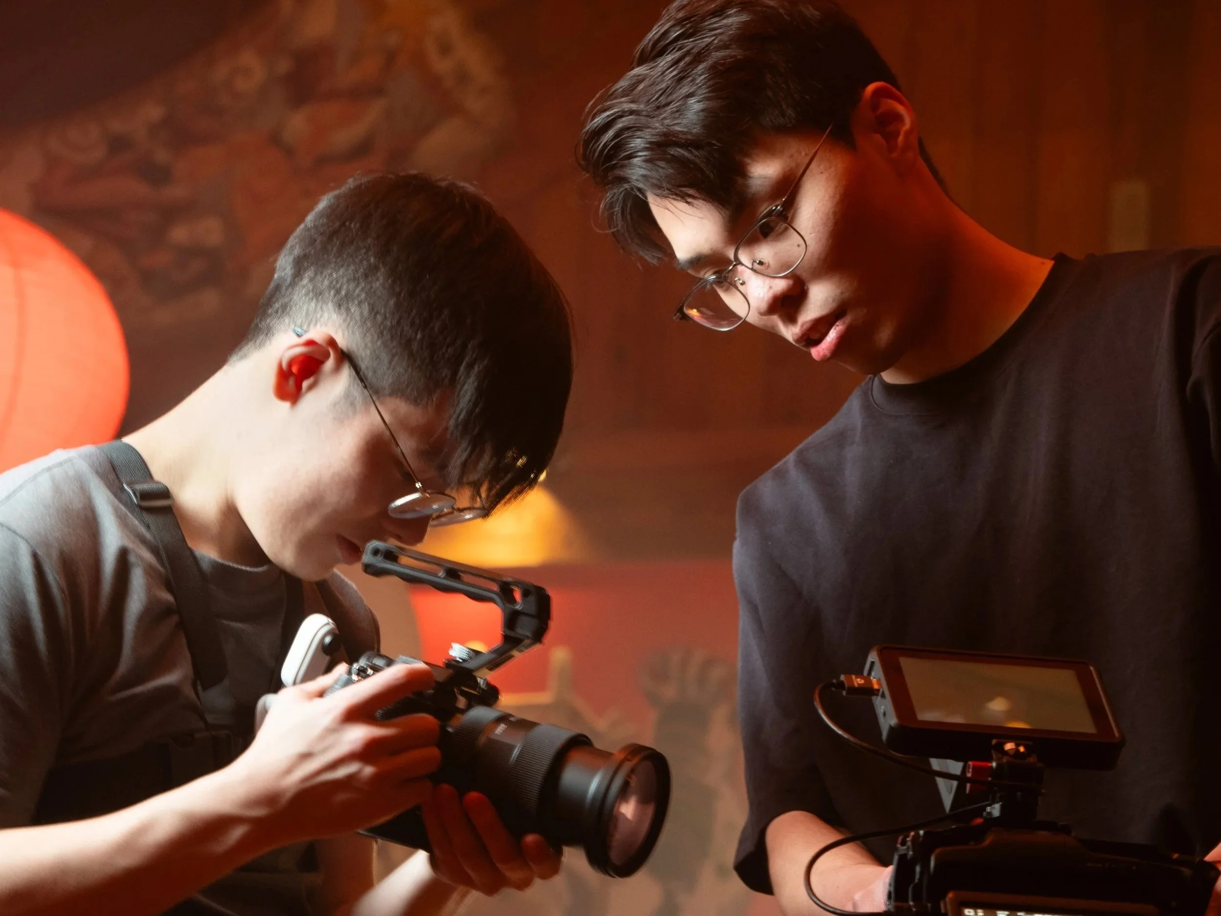 Two young men, both wearing glasses, are closely examining a camera and a device with a screen, possibly during a photo shoot or video production in a warmly lit room.