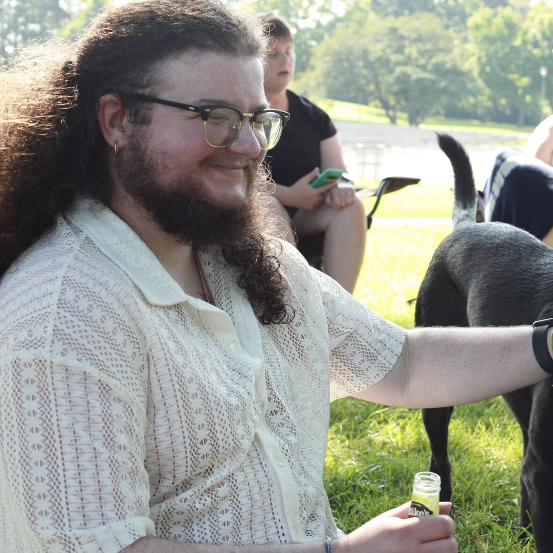 Profile shot of Bernie Ludi, a Romani man with long curly black-brown hair and a curly beard. They are outside, gleefully petting a black dog. They have browline glasses, red hearing aids, and wear a cream lace shirt.