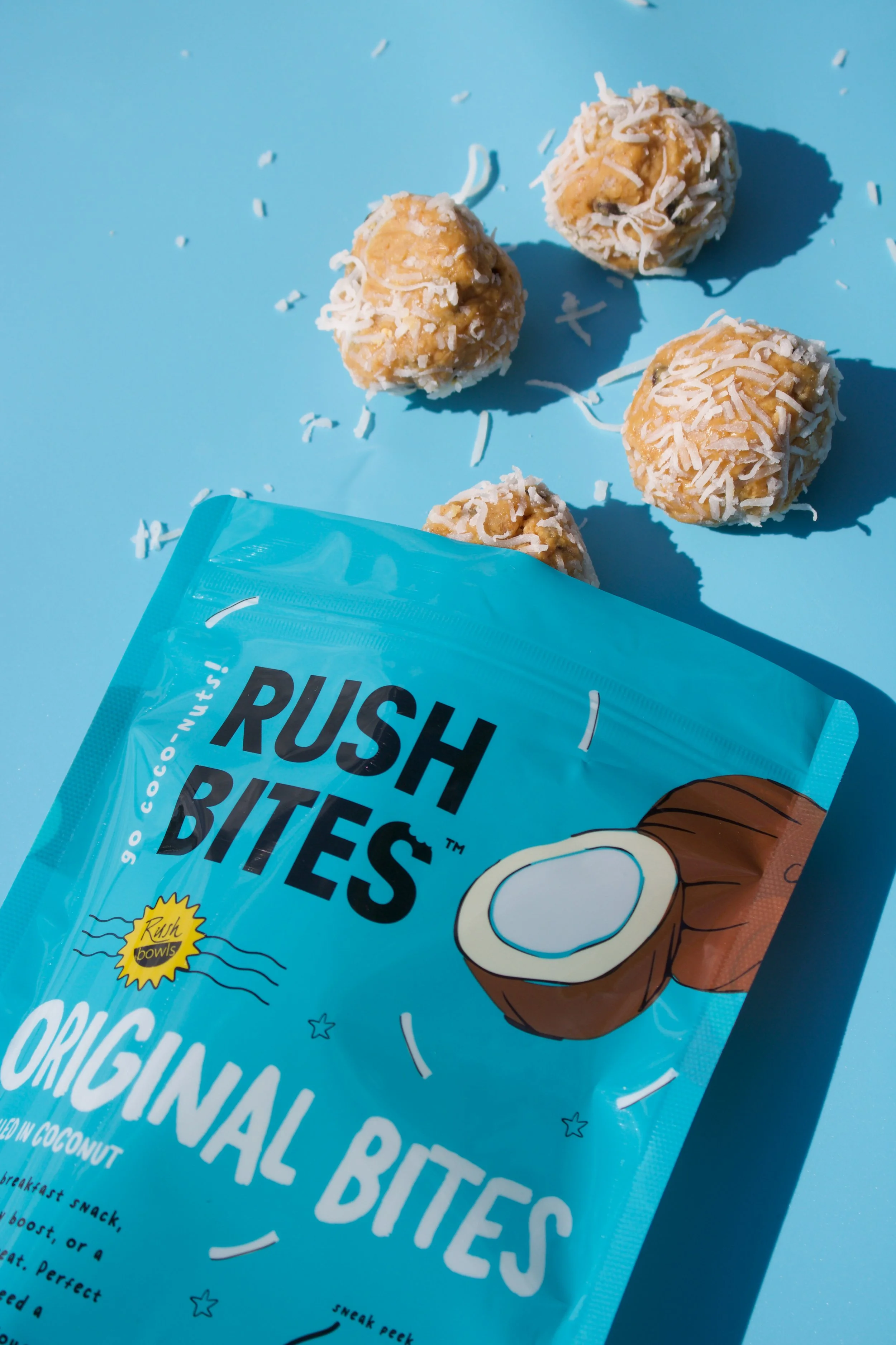 A bright blue package of Rush Bites coconut-flavored snack balls with shredded coconut on top, placed on a light blue surface. Several snack balls are scattered nearby.