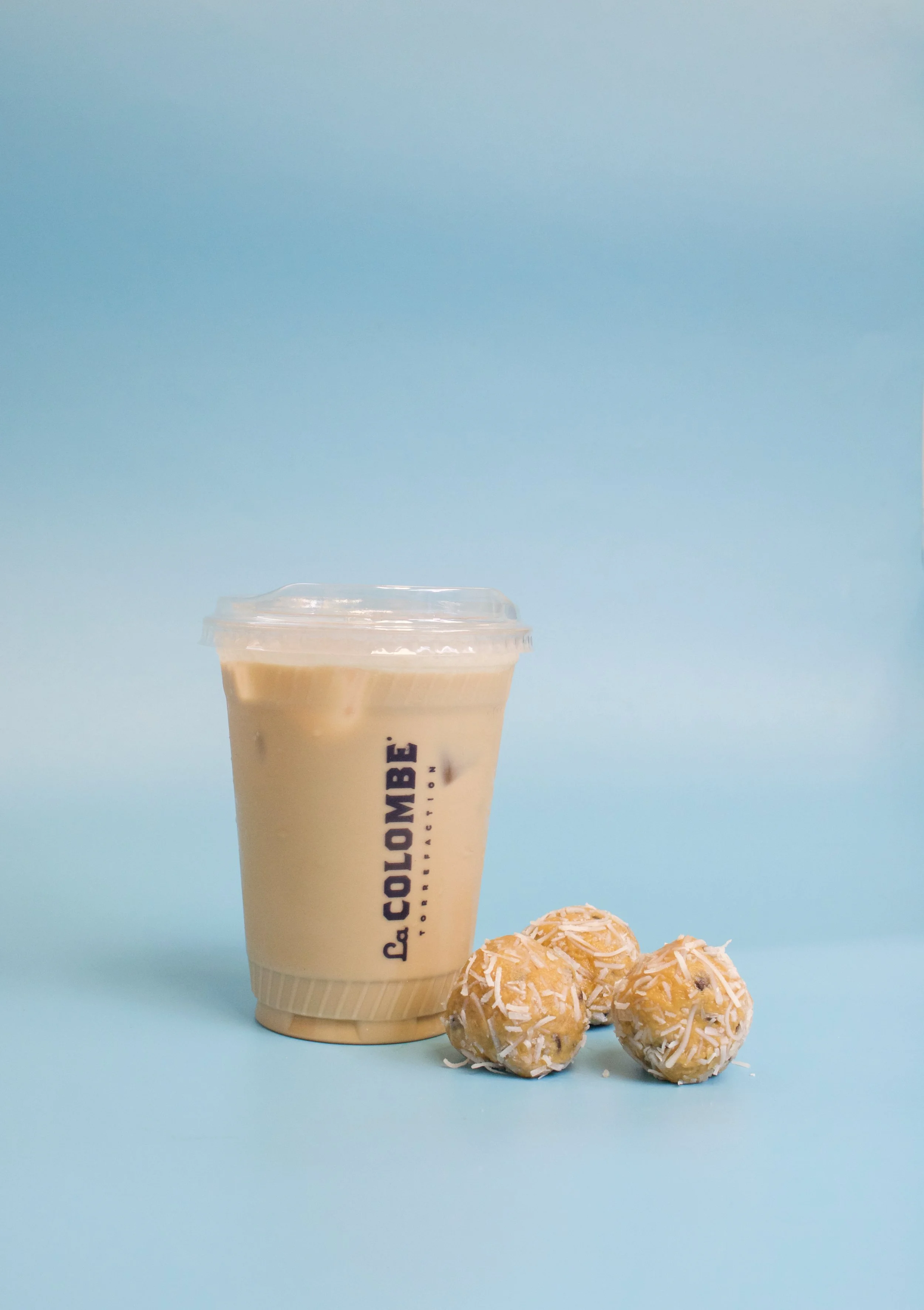 A plastic cup of iced coffee with a lid, labeled 'L. COLOMBE BAKERY,' placed next to three coconut-flavored energy bites coated with shredded coconut, on a light blue background.
