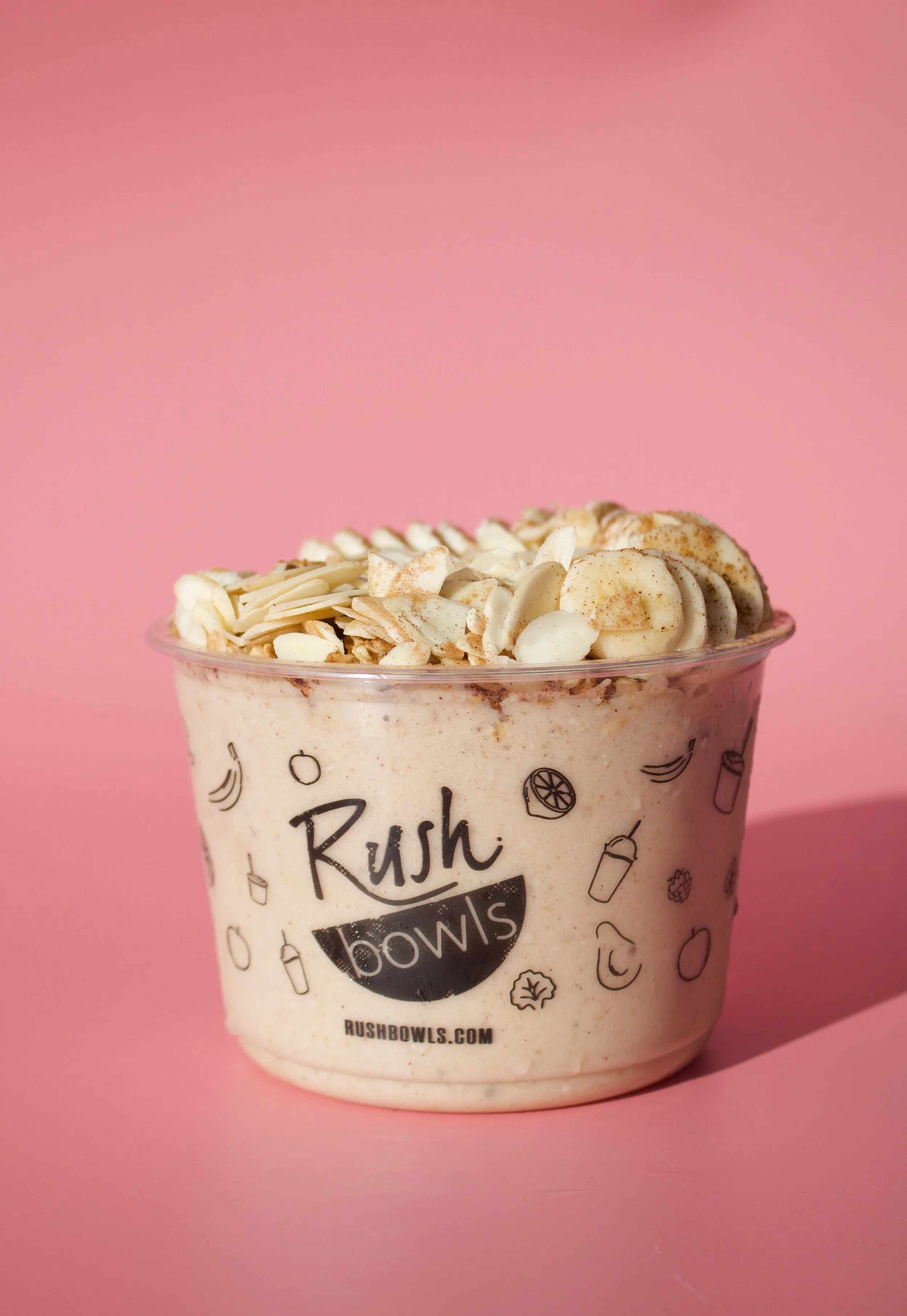 A cup of frozen yogurt topped with sliced almonds and cinnamon, labeled with the Rush Bowls logo and placed against a pink background.