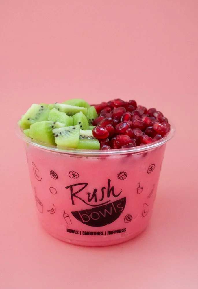 A clear plastic bowl filled with sliced kiwi and pomegranate seeds against a pink background. The bowl has black and pink text that says "Rush bowls" and mentions bowls, smoothies, and happiness.