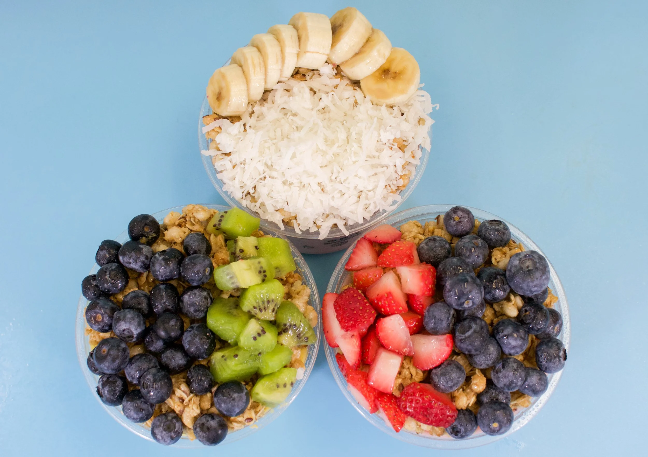 Three bowls of granola parfaits topped with blueberries, strawberries, sliced bananas, and kiwi on a light blue background.