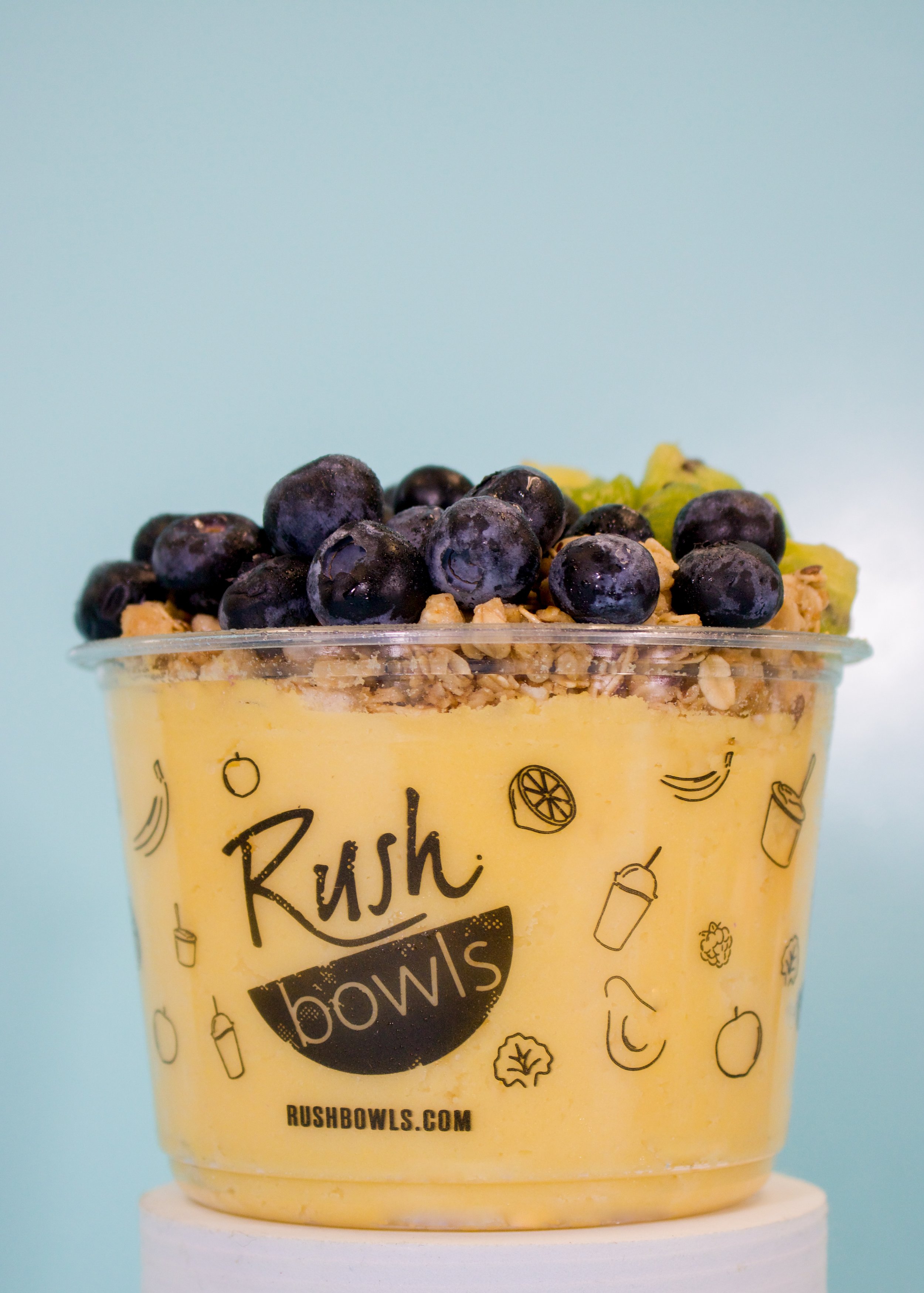 A clear plastic container filled with a yellow smoothie topped with blueberries, granola, and green fruit. The container has a black logo with the words 'Rush Bowls' and small illustrations of fruits and drinks.