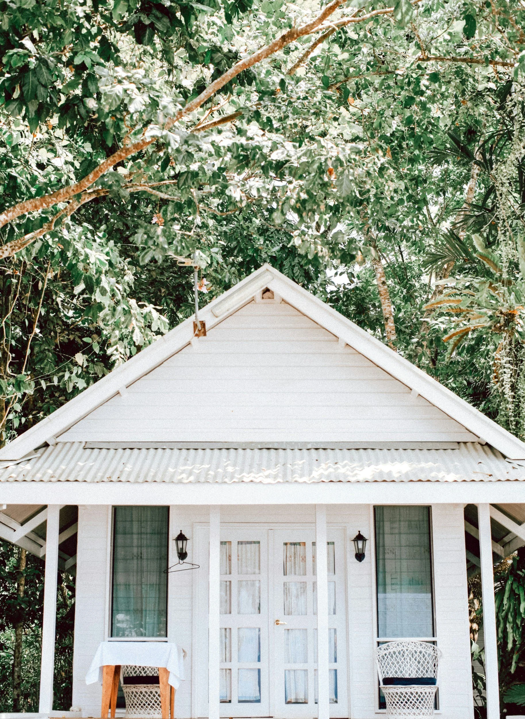 A small white cottage with a gabled roof, flanked by two windows with curtains, and a door, situated in a lush, green garden with dense foliage, symbolizing rest, relaxation, and a safe haven.