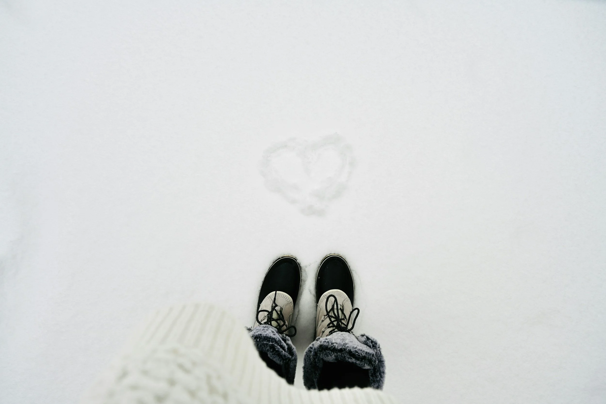 Women stands in snow in winter boots, jeans, and a sweater with a hand-drawn heart in the snow representing self-care and self-compassion as mental health tips for the holidays including Christmas, Hanukah and New Year's.