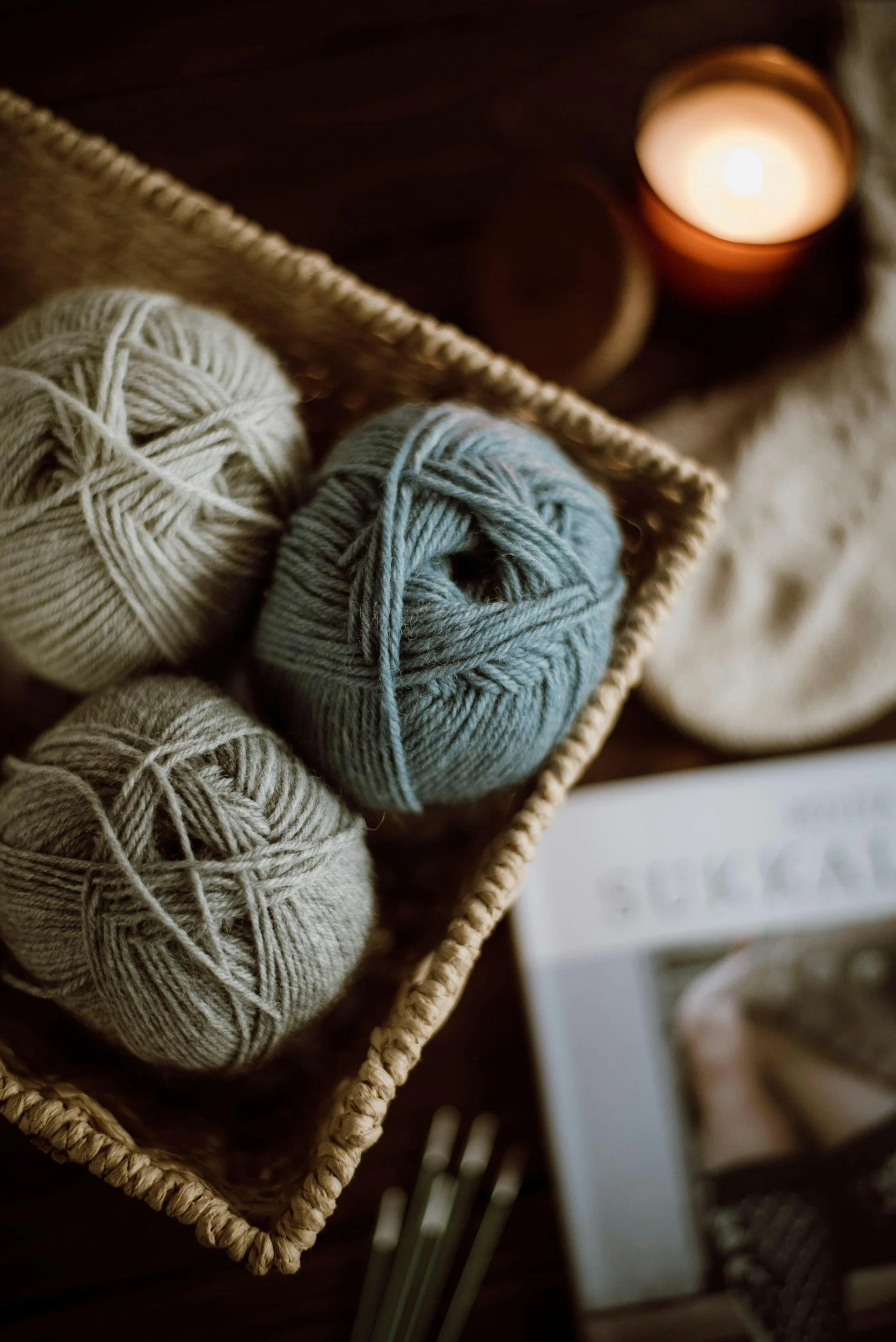 Neutral colored balls of yarn in a basket on a table with a magazine and lit candle, indicting the soothing and relaxing comfort of cozy analog hobbies like knitting and crocheting.