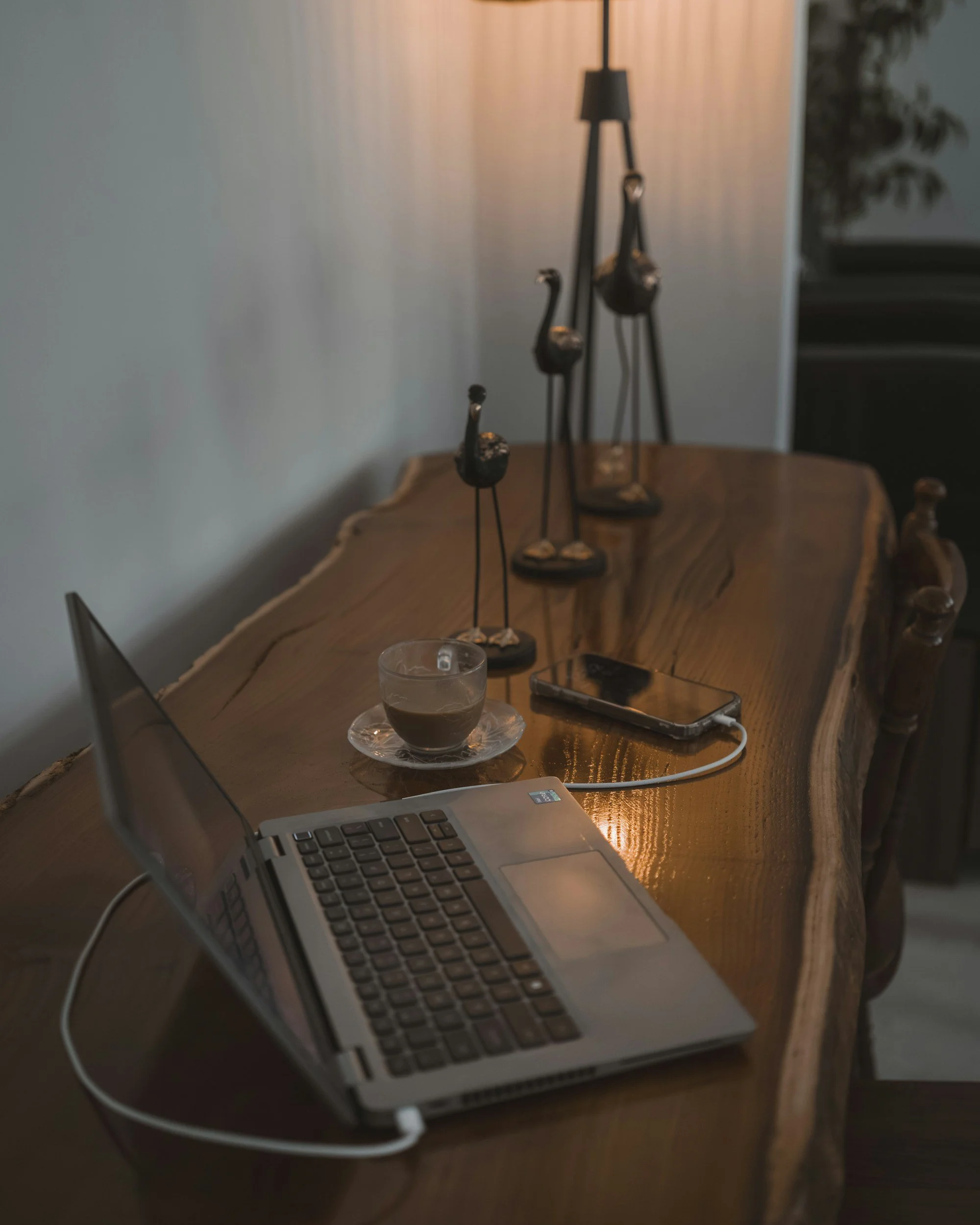 A wooden table with a laptop, a cup of coffee, a smartphone connected to a charger, and decorative sculpture of three cranes. Representing holistic online therapy from home.