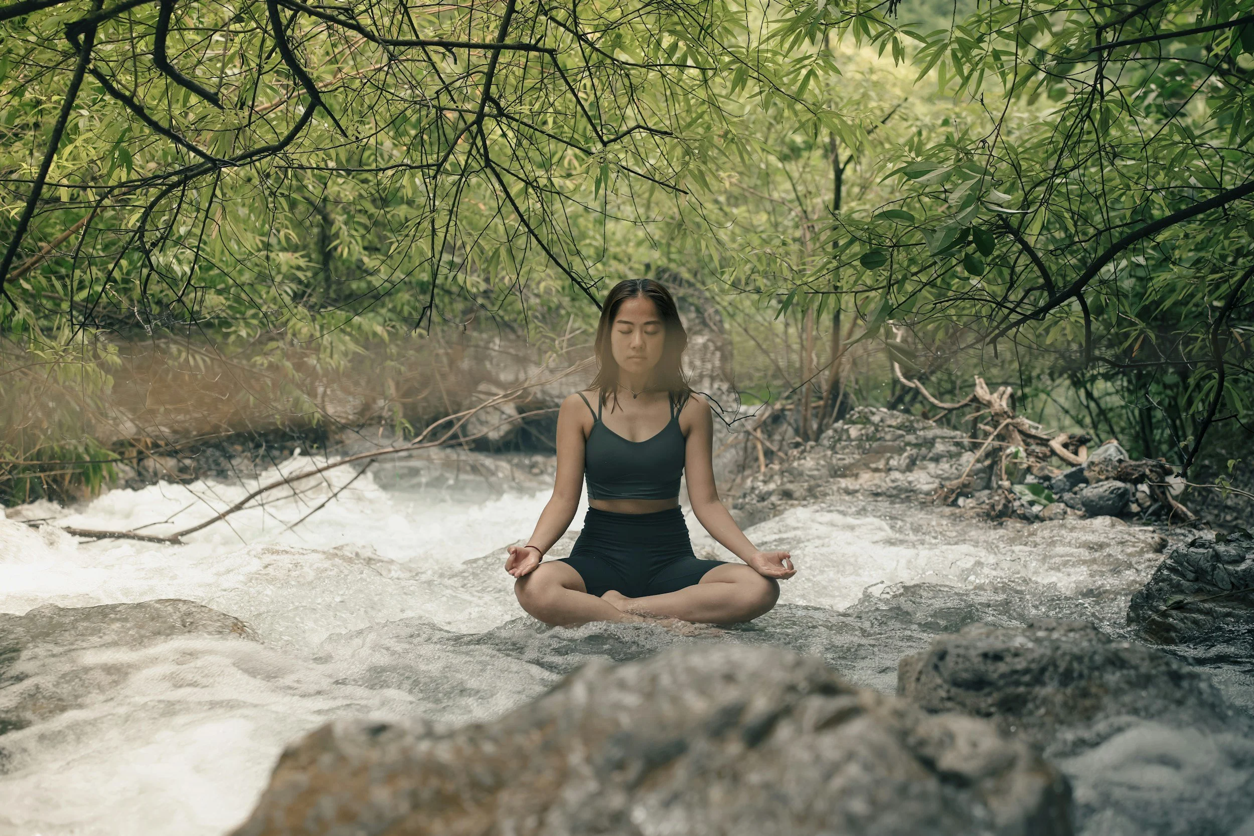 A woman meditating in a seated lotus position on a rock in a river, surrounded by lush green trees and branches overhead, with steam rising from the water.